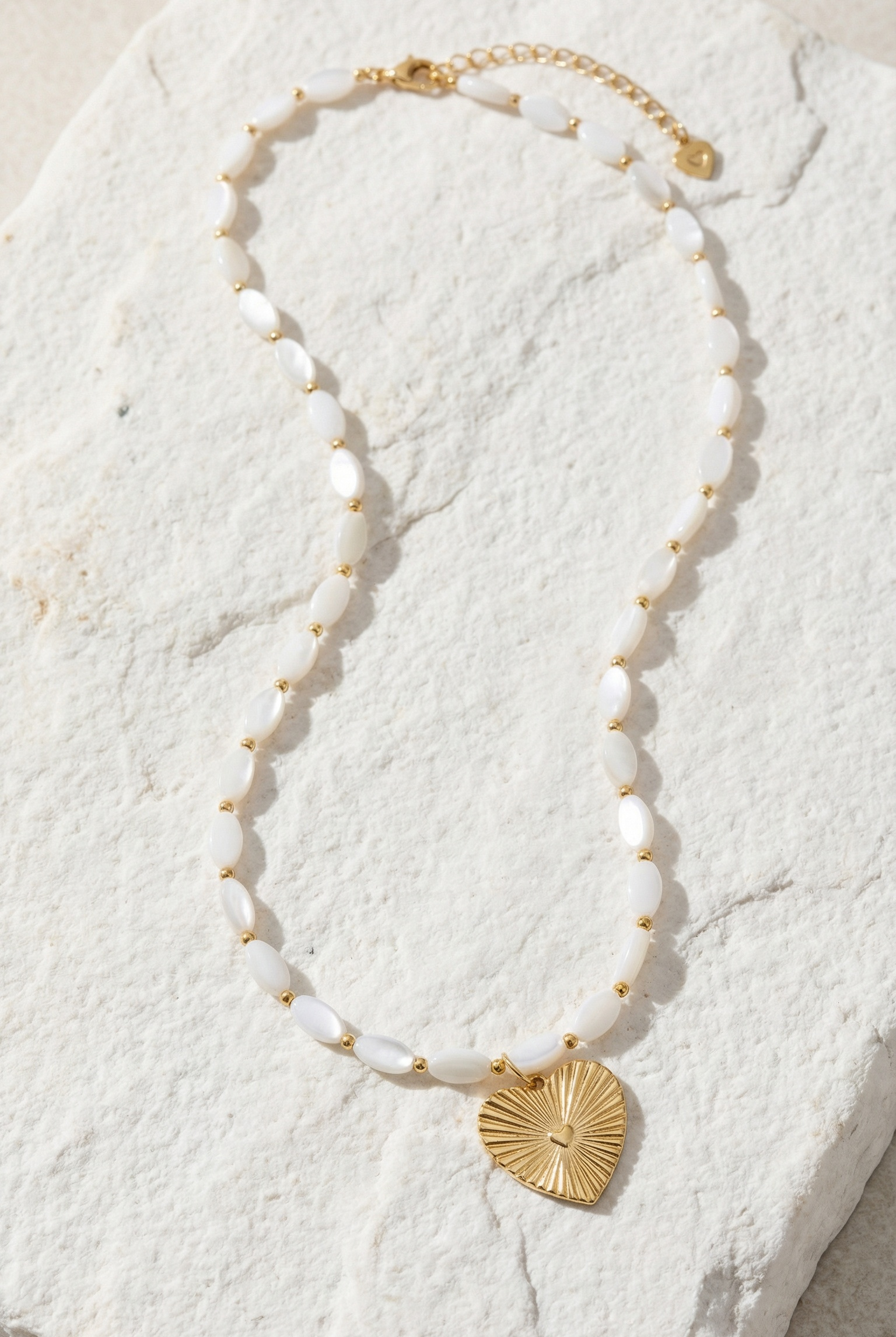 Necklace with white mother pearl beads and gold heart pendant on a textured white surface