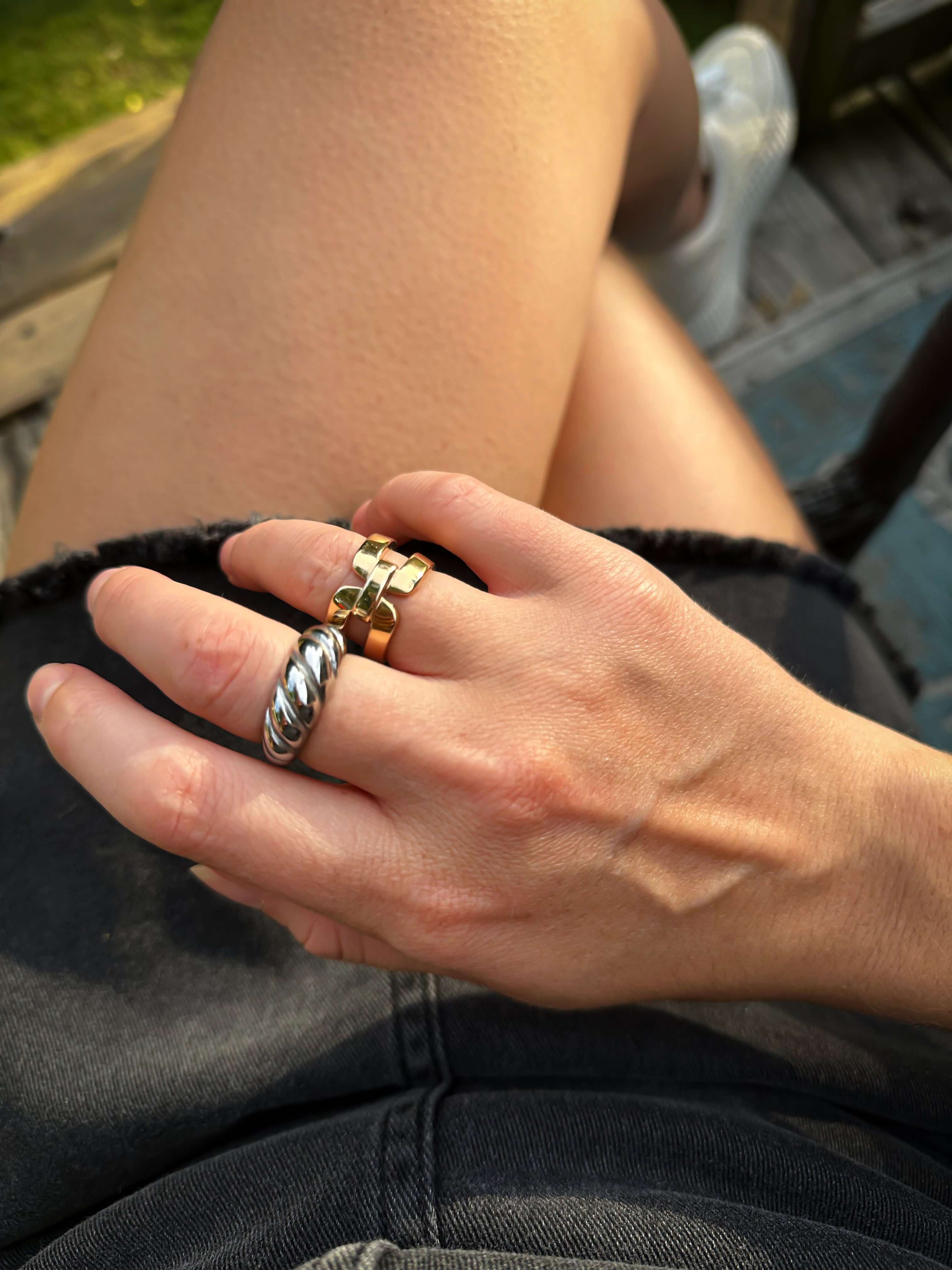 Close-up of a hand adorned with the Croissant Ring and silver bands in domed settings, resting on bare legs clad in black denim shorts.