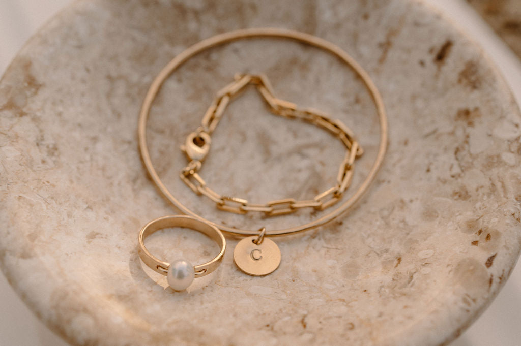 Gold ring, bracelet, and chain necklace with a 'C' pendant, displayed on a beige marble dish.