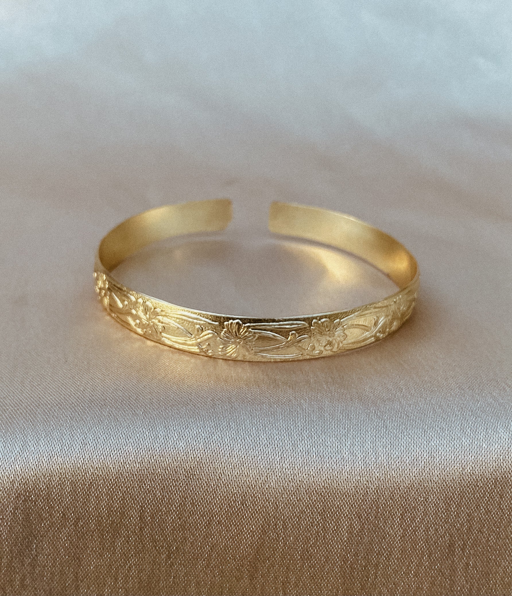 A gold cuff bracelet with intricate floral engravings, displayed on a soft beige fabric backdrop.