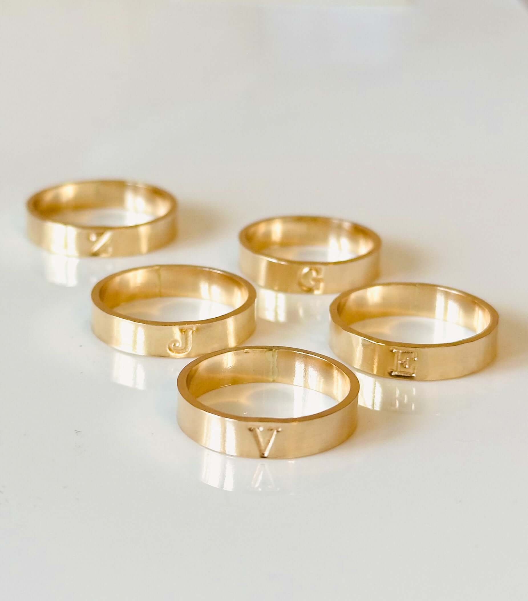 Five Initial Band rings made of 14k gold-filled, each engraved with the letters C, J, E, V, and G, arranged on a white surface.