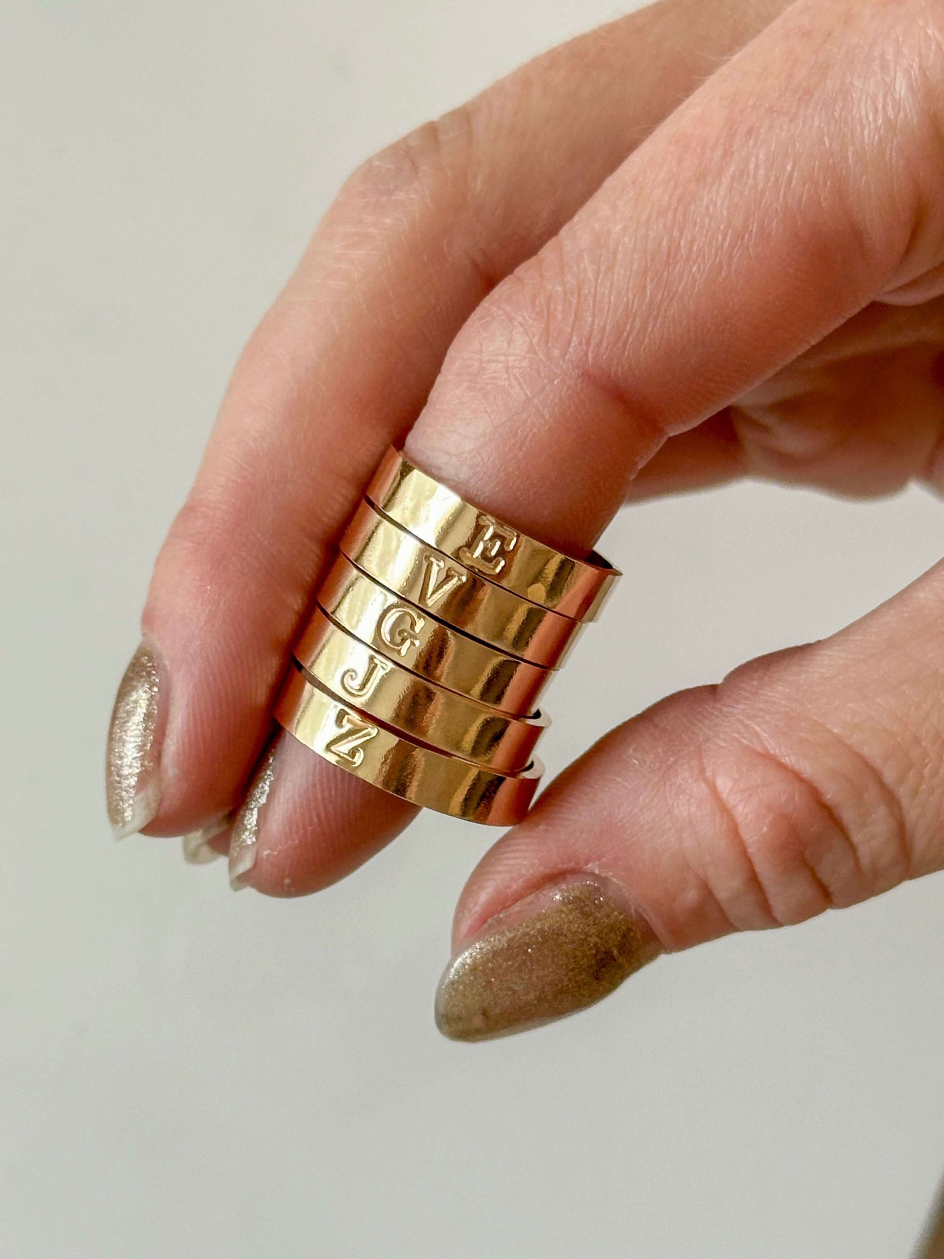 A hand adorned with gold, glittery nail polish displays several stacked Initial Bands on its fingers, each engraved with a letter and crafted from 14k gold-filled material.