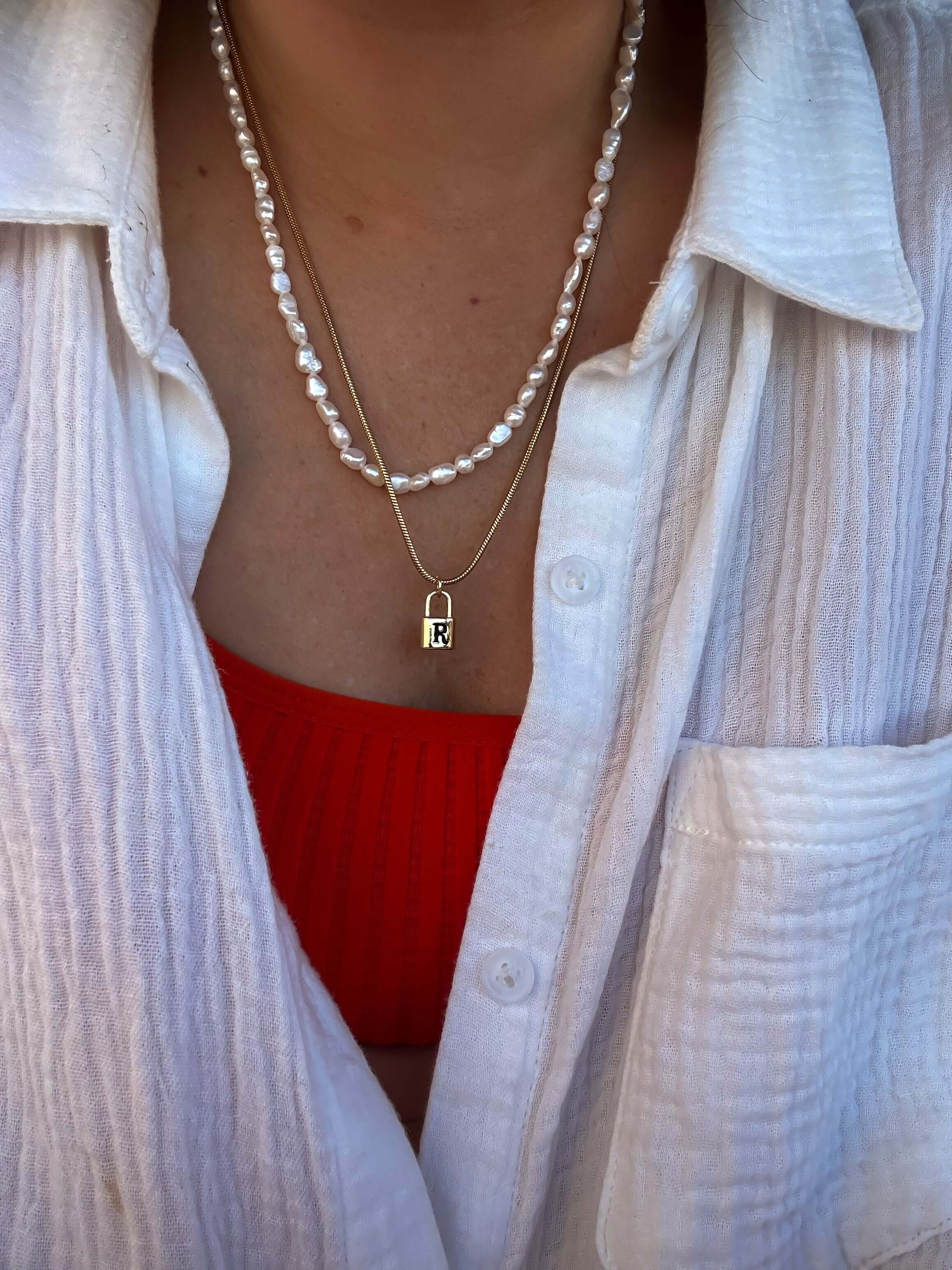 Person wearing a white button-up shirt, a red top, and two necklaces: one with pearls and one featuring the Lock Initial Necklace for added personalization.