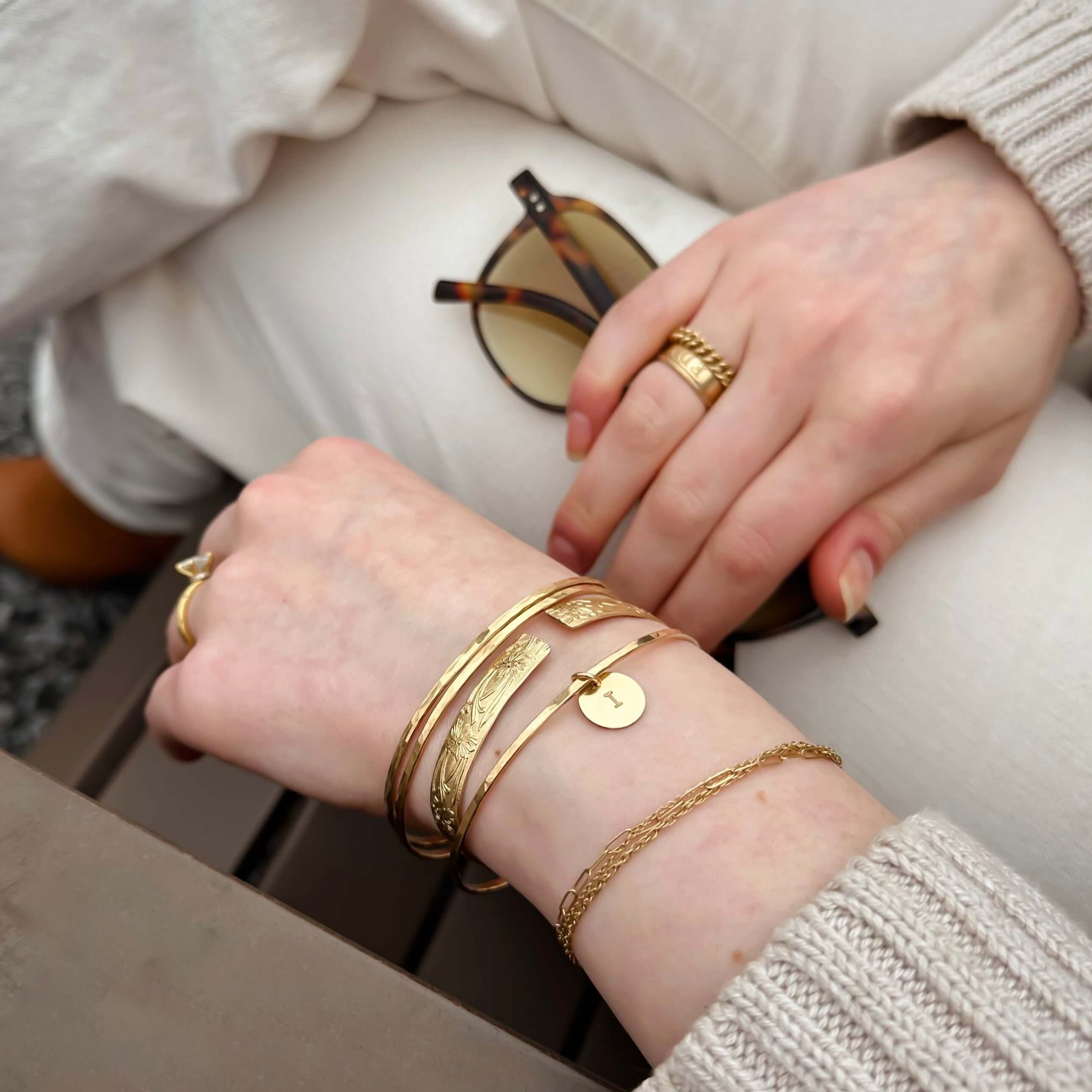 Close-up of person’s hands featuring a Marigold Cuff Bracelet, gold rings, and holding sunglasses. They are dressed in light-colored clothing decorated with floral details.