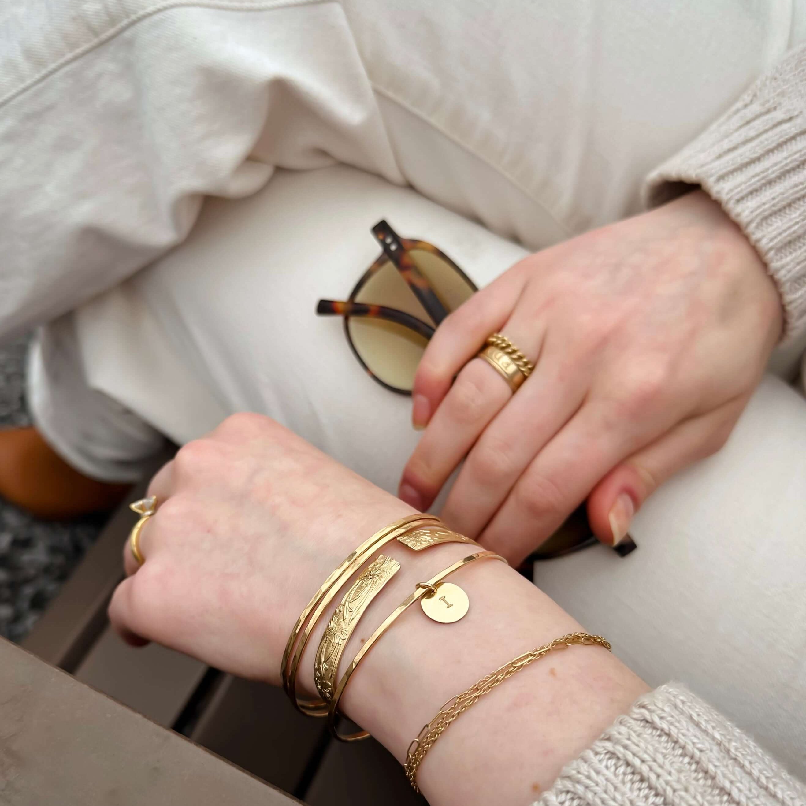 Hands adorned with gold bracelets and rings rest on white-clad legs, holding sunglasses, with a grey sweater sleeve visible, featuring the Initial Bangle for a touch of personalization.