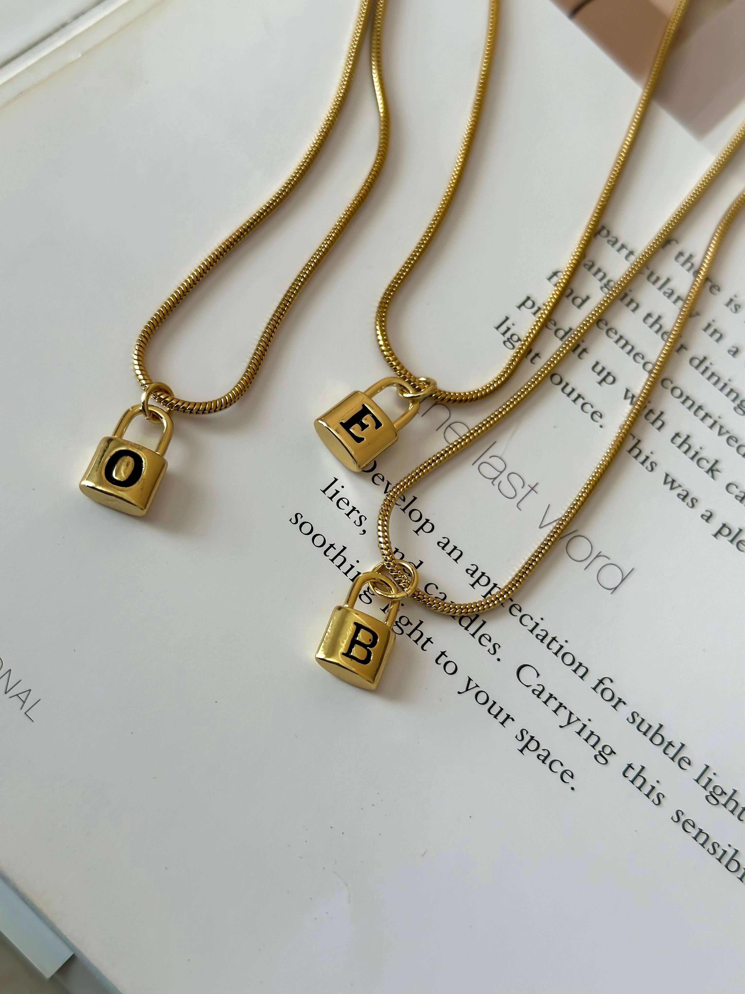 Three Lock Initial Necklaces, personalized with lock pendants featuring the letters "O", "E", and "B," are elegantly displayed on an open book and draped on delicate snake chains.