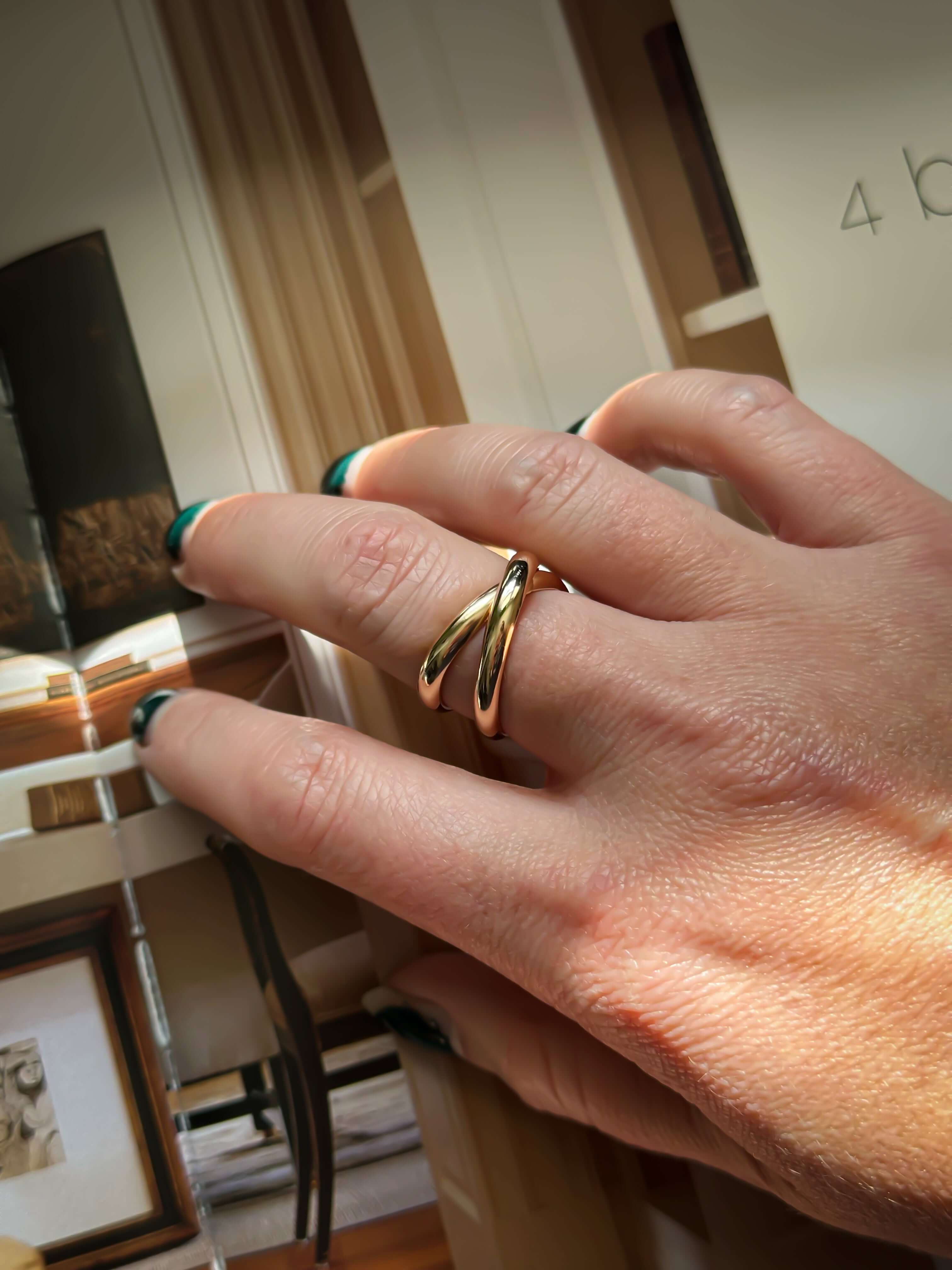 A hand adorned with the Crossover Ring in gold, featuring two elegant loops, is resting on an open book and showcasing beautifully teal-painted nails.