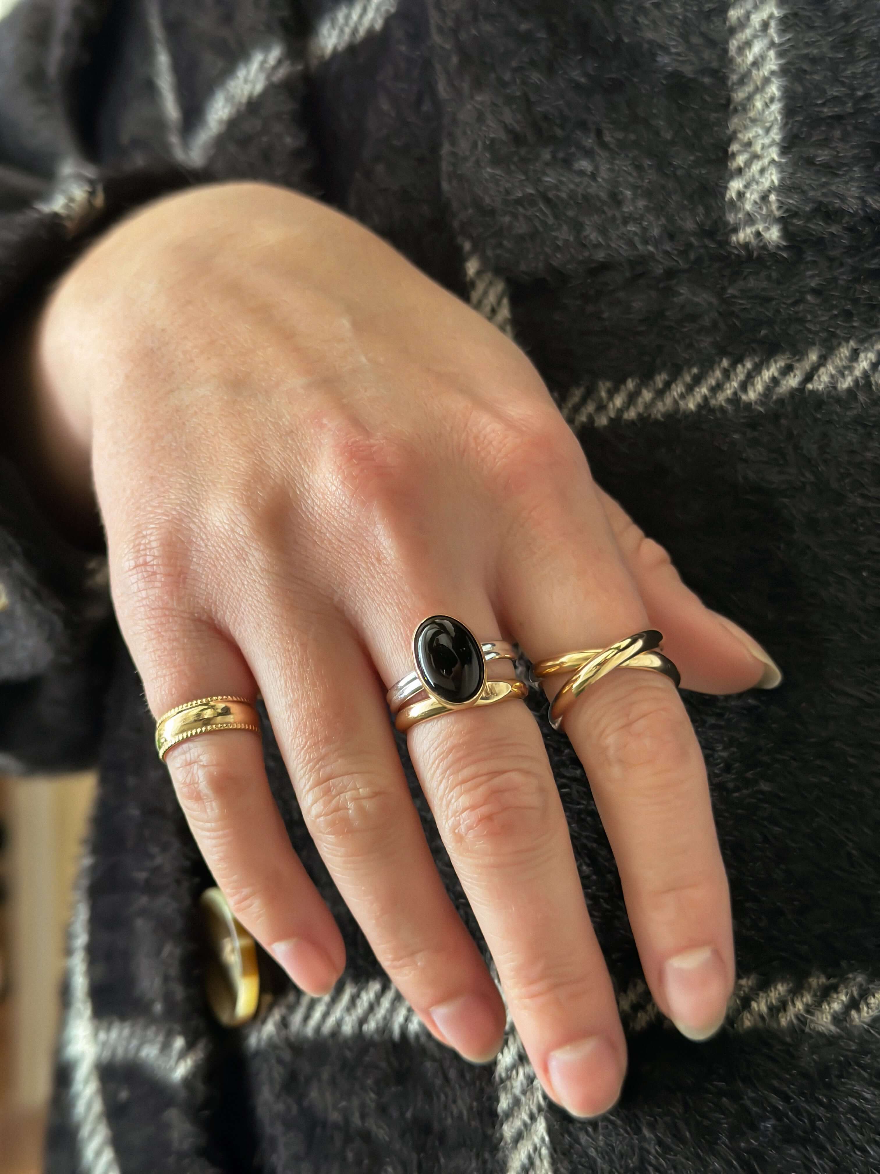 Close-up of a hand adorned with multiple rings, prominently featuring the Crossover Ring with a black stone, showcasing trendy jewelry with a chunky look.