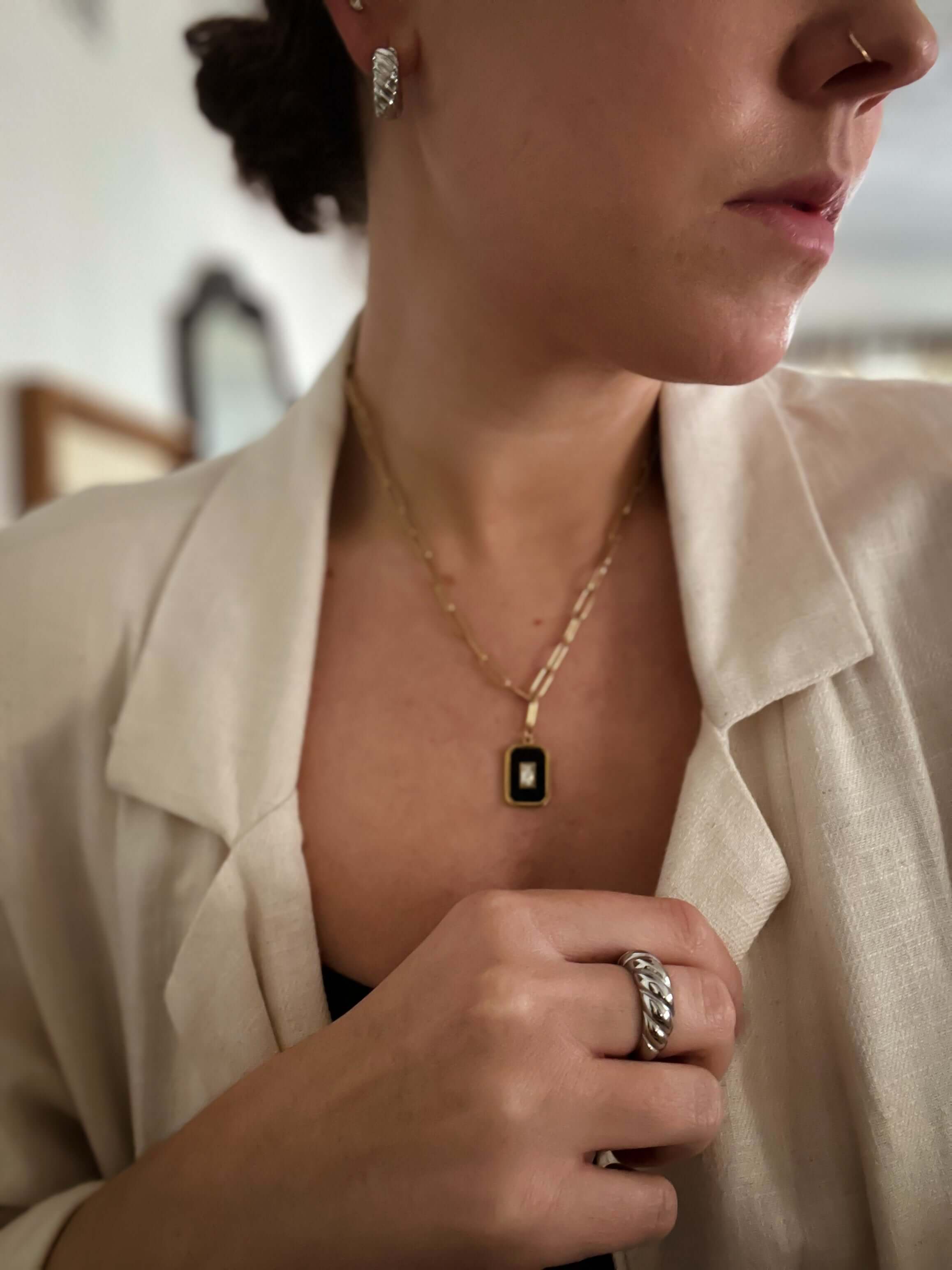 Close-up of a person wearing a light-colored blazer, an Eve Necklace featuring a black hexagon pendant, along with matching earrings and a ring on their finger.