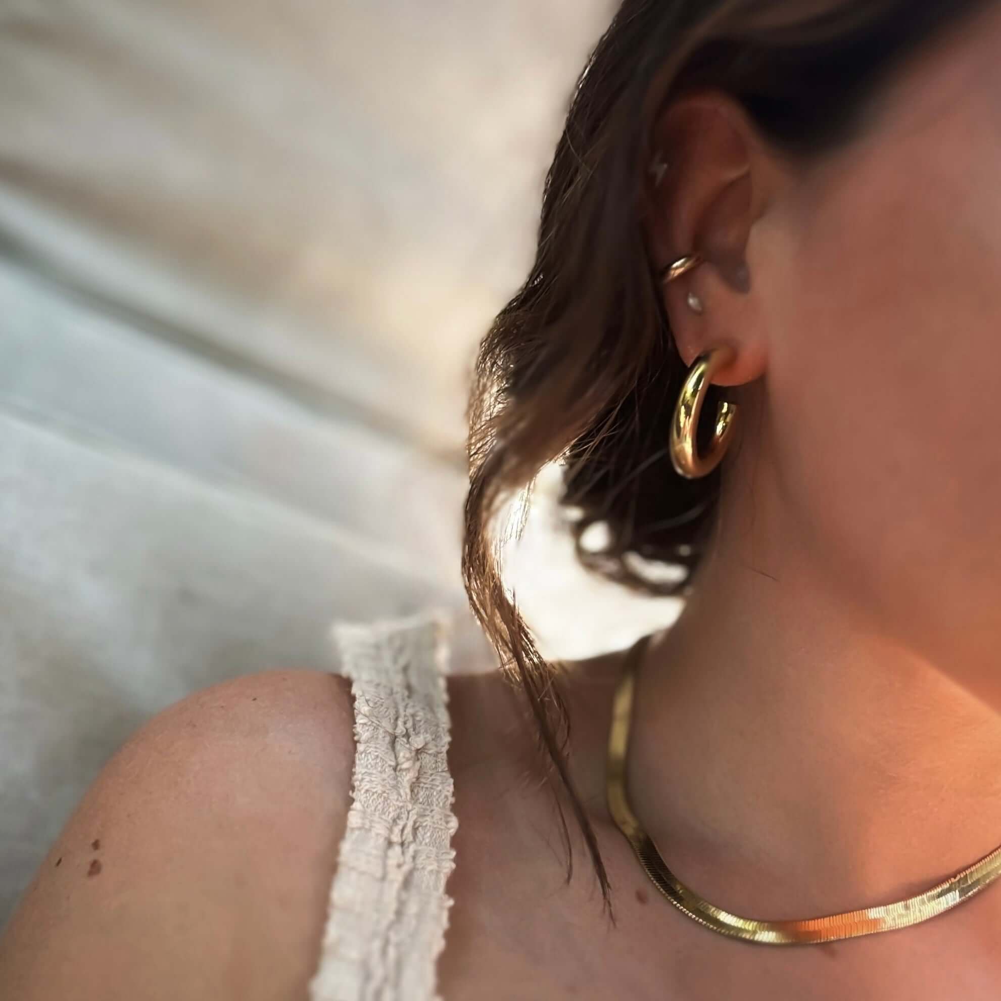 Close-up of a woman's shoulder and neck adorned with Cali Tube Hoops and a gold necklace, wearing a white sleeveless top.