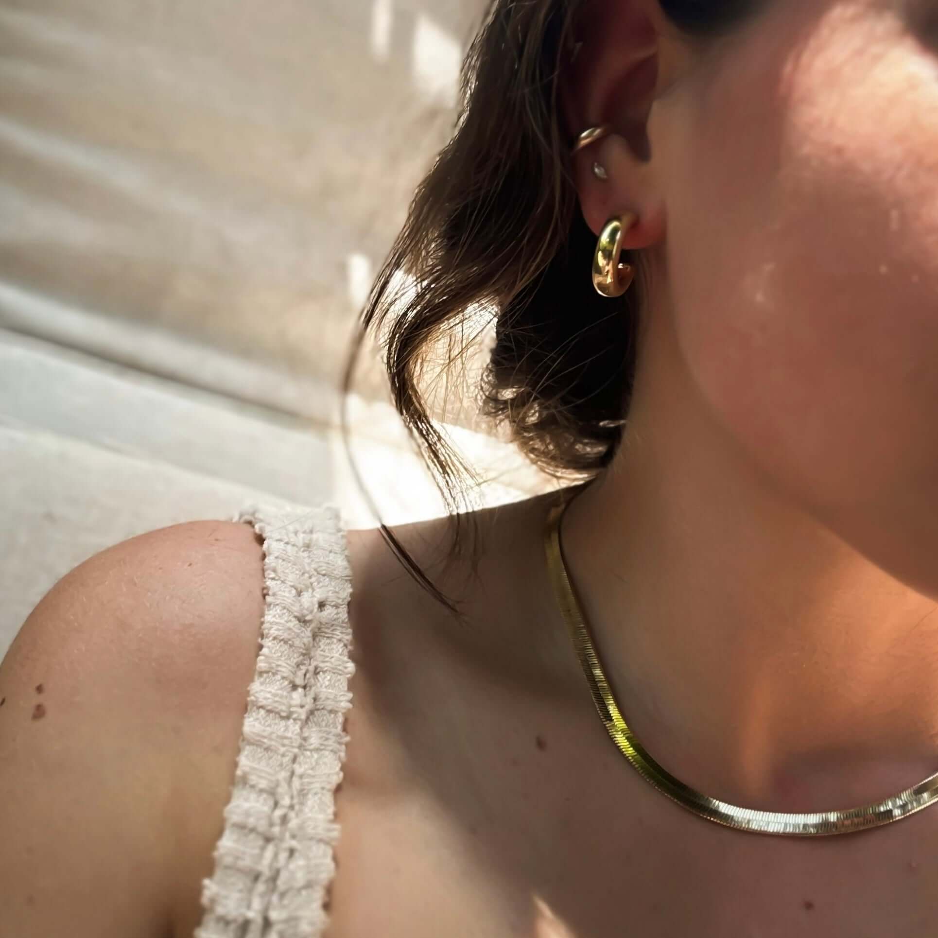 Close-up of a woman's neck and shoulder wearing a tank top, Cali Tube Hoops earrings, and a matching gold necklace.