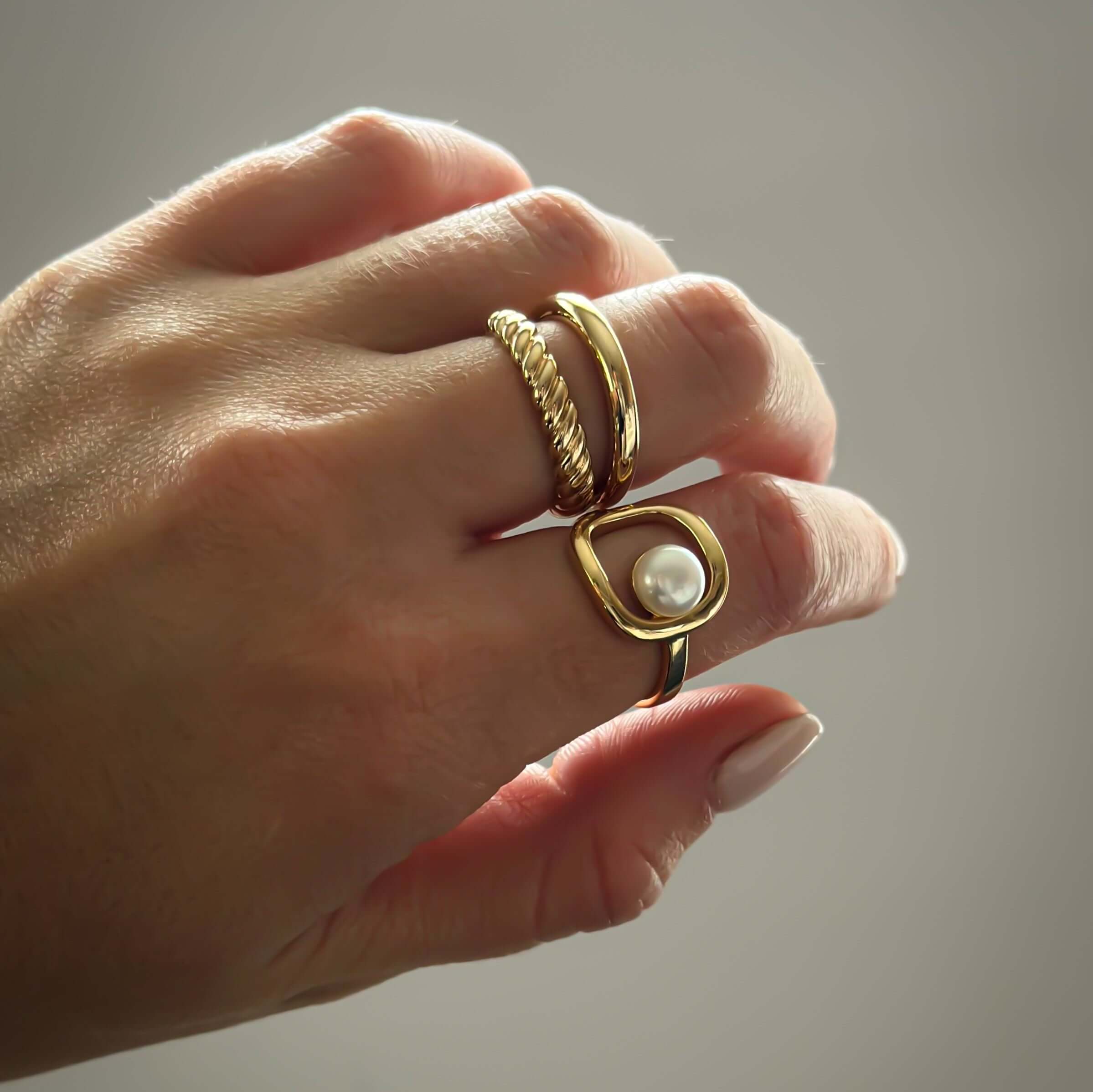 Close-up of a hand wearing three rings, including an elegant and bold gold ring with a pearl and the Diana Double Ring from Apple & Figs, against a plain background.