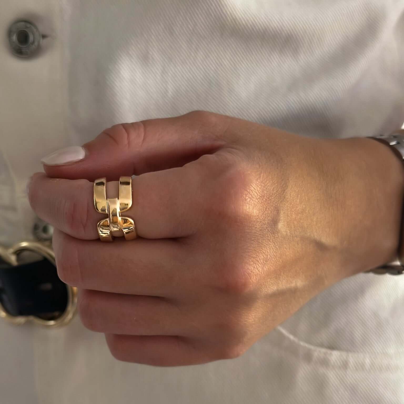 Close-up of a person's hand wearing the Double Buckle Ring with an 18k gold plating and a unique design, resting against a white shirt with a black belt.