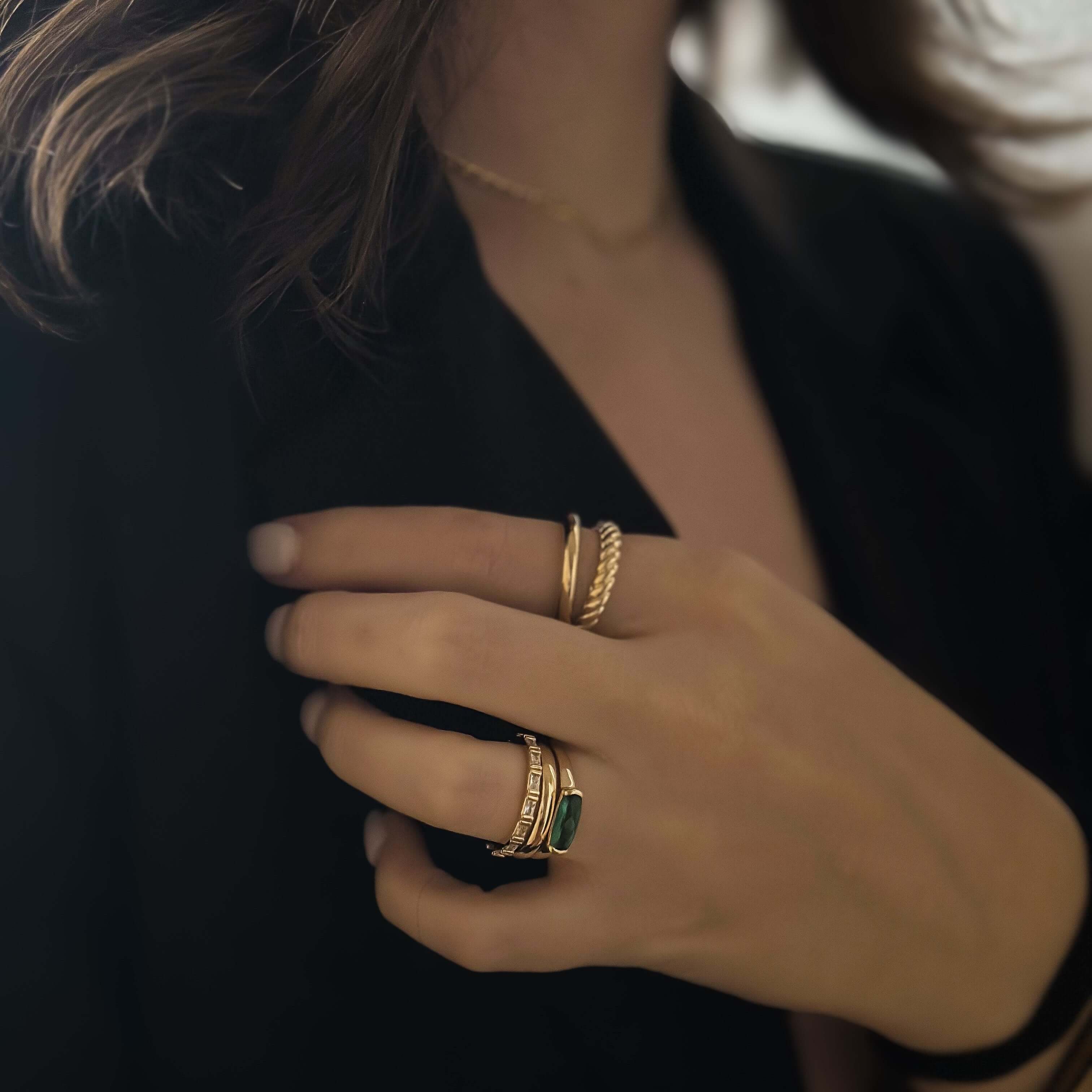 A person wearing a black top showcases their hand adorned with multiple gold rings, including the Emerald Stacker Ring which features a radiant green stone, accompanied by a necklace glinting in 18k gold plating.