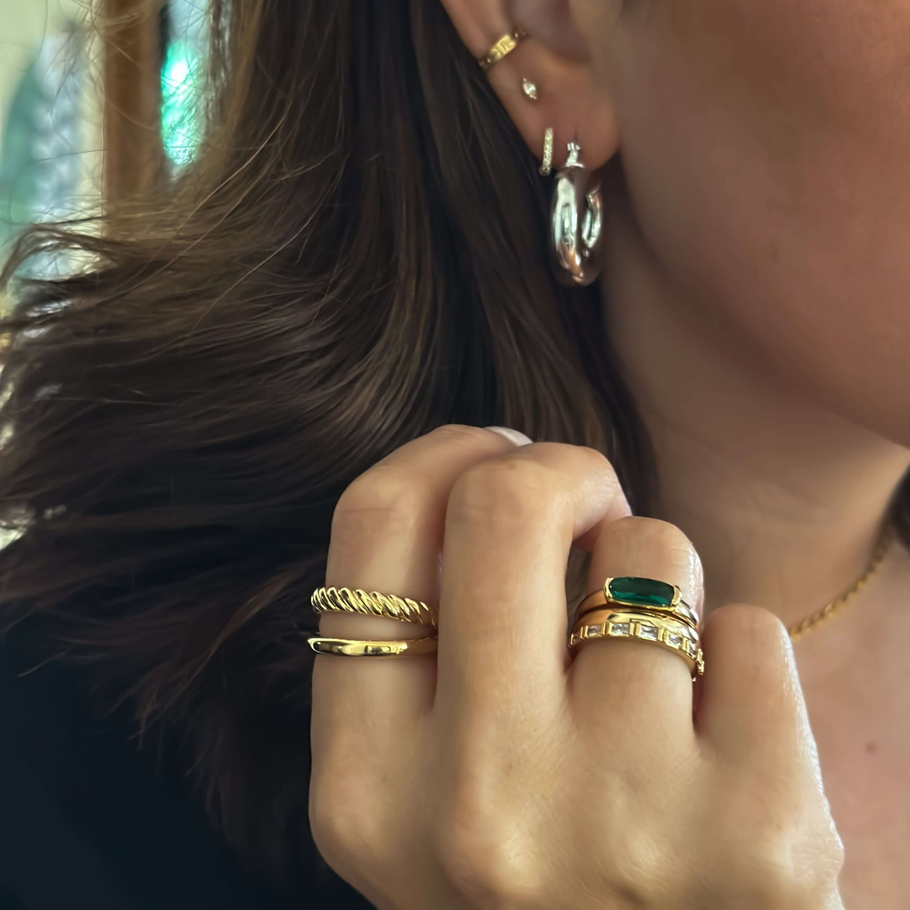 Close-up of a person adorned with multiple gold rings and earrings, prominently featuring the stunning Emerald Stacker Ring with 18k gold plating, as they hold their hand near their face.