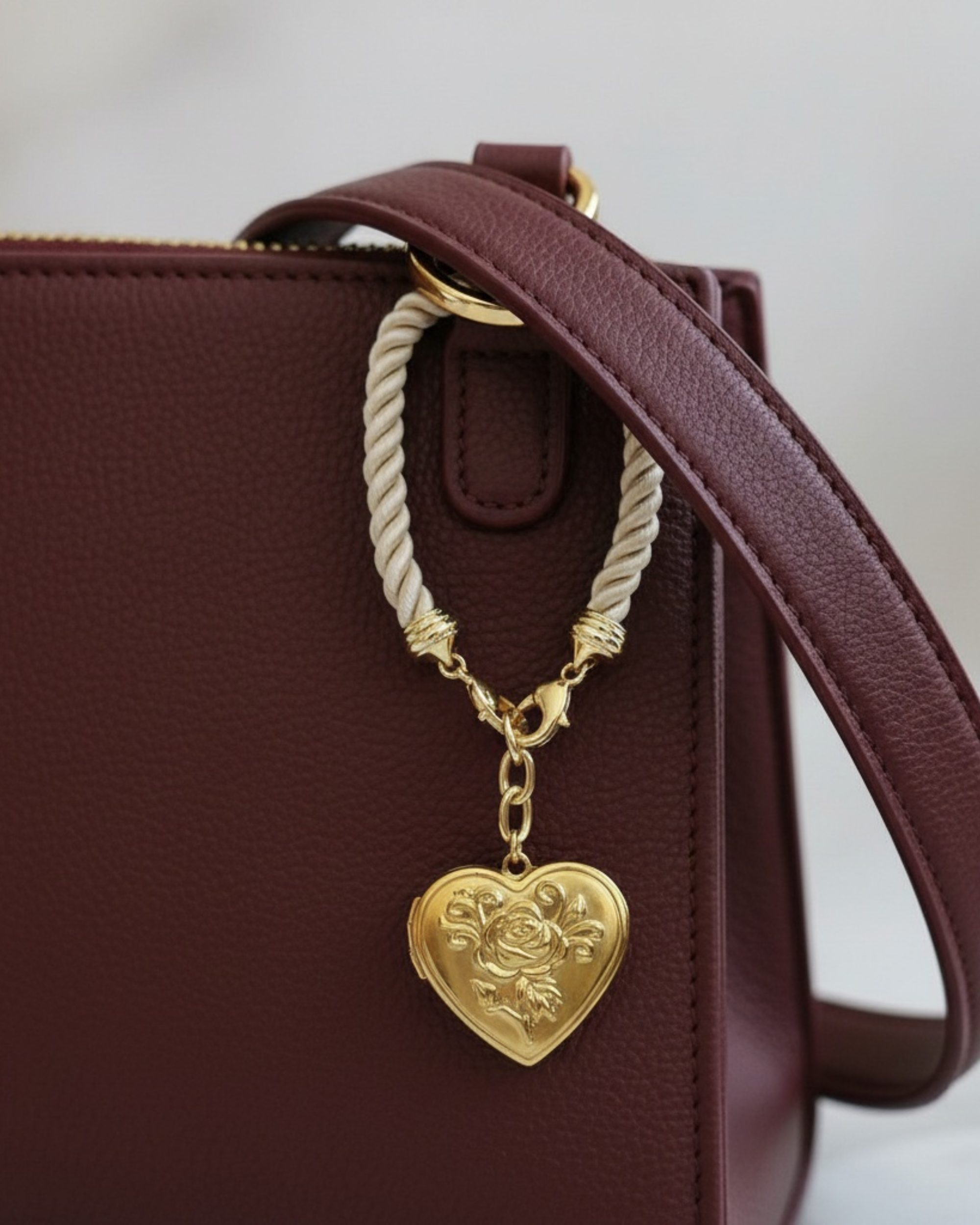 Gold heart-shaped locket bag charm with engraved floral detail, suspended from a cream twisted cord and brass ring