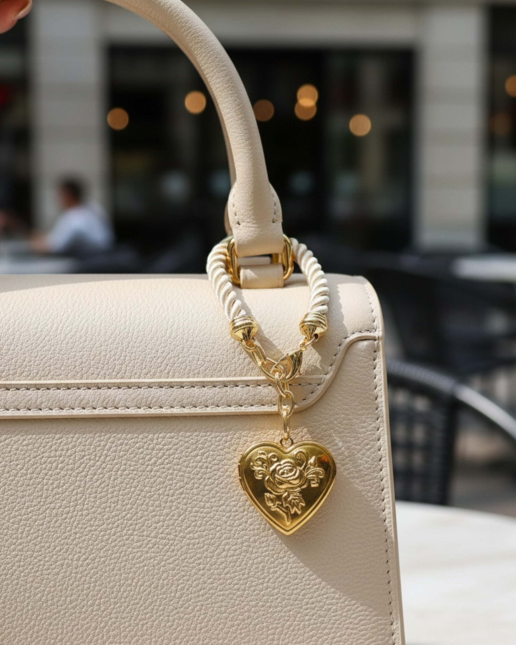Gold heart-shaped locket bag charm with engraved floral detail, suspended from a cream twisted cord and brass ring
