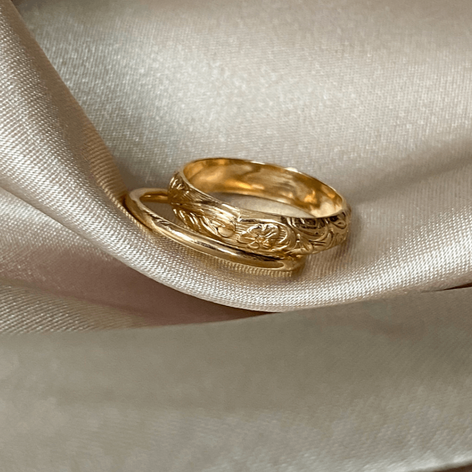 The Floral Ring Set, featuring two gold bands with intricate designs, rests on a soft, textured, cream-colored fabric.