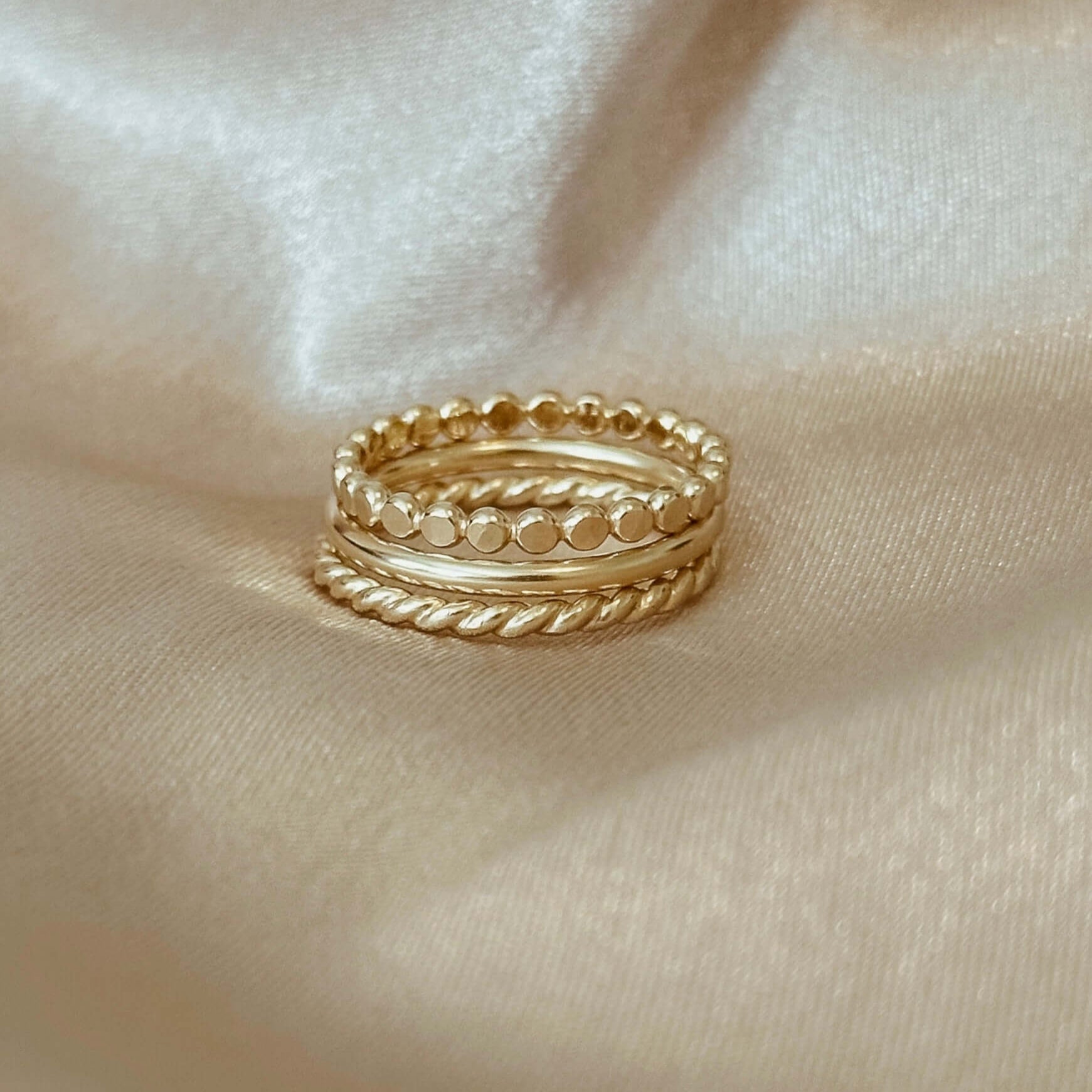 The Stacking Ring Set, featuring three 14k gold filled rings with varying textures—including a smooth band—rests elegantly on a silky beige fabric backdrop.