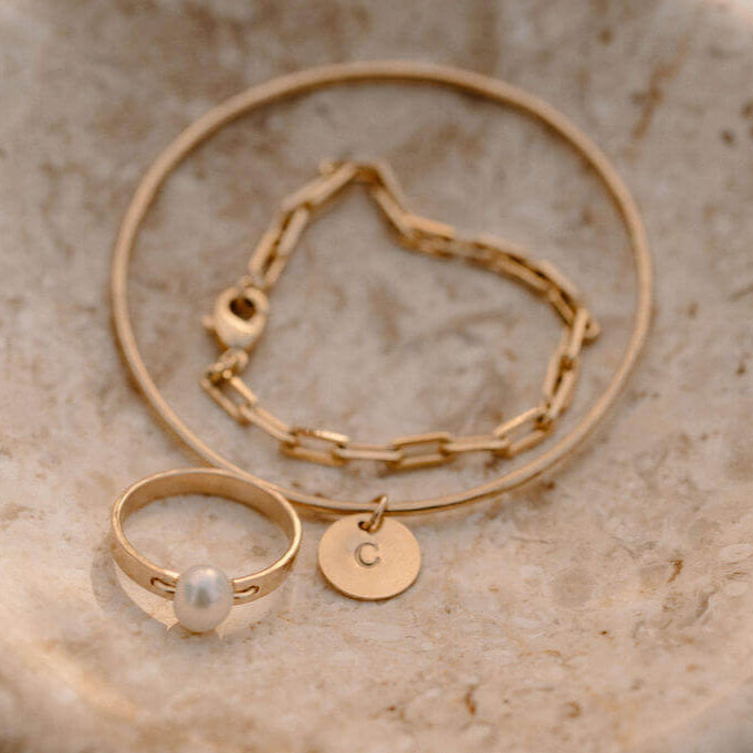 Initial Bangle, chain bracelet with an initial charm pendant, and a pearl ring on a marble surface.