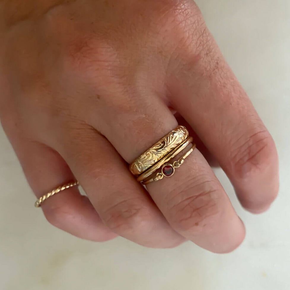 A hand adorned with the Floral Ring Set, featuring a textured gold band, a twisted gold band, and a vintage floral charm ring with a small red gem.