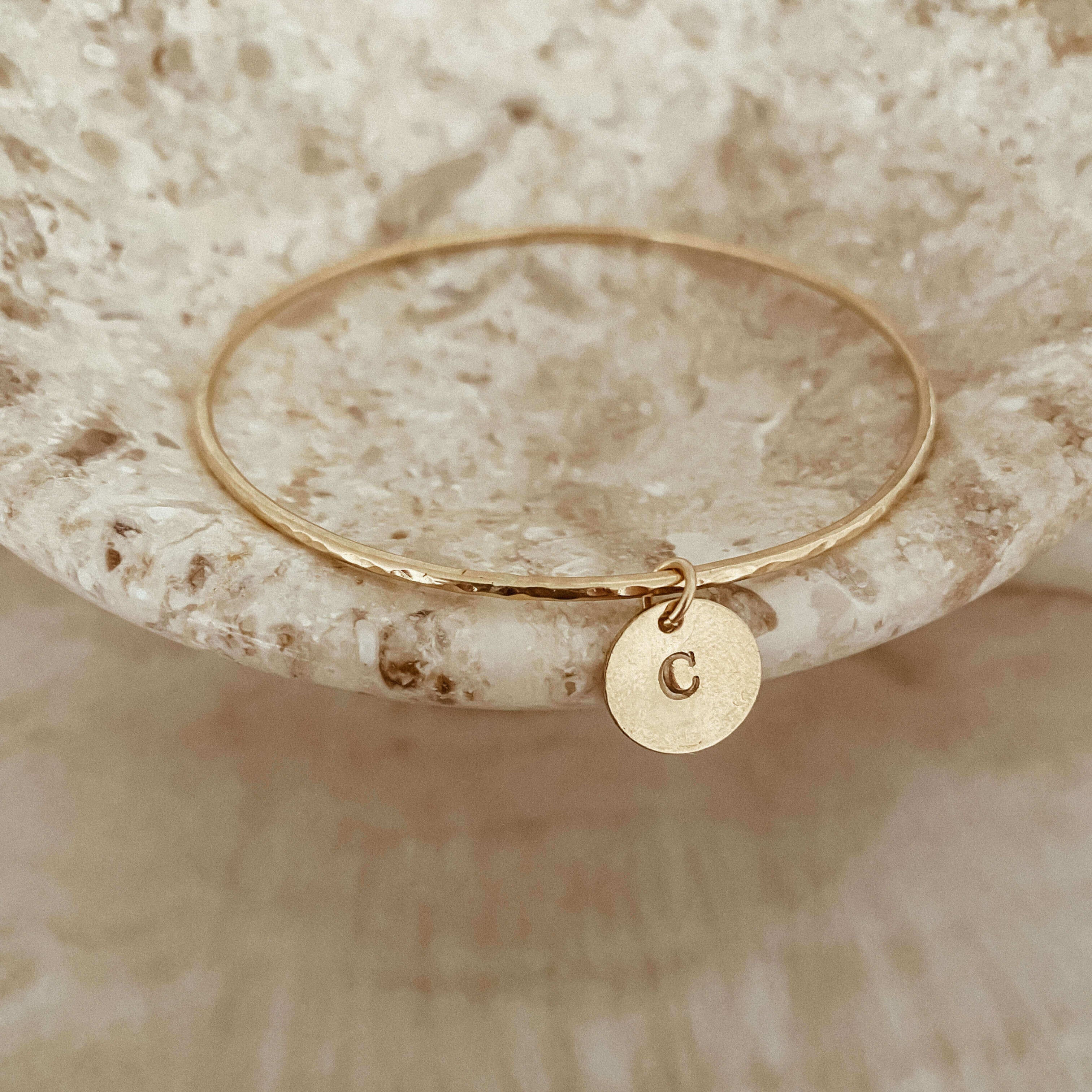 The Initial Bangle displayed on a textured stone surface showcases a small, hand-stamped circular pendant featuring an initial charm engraved with the letter "C".