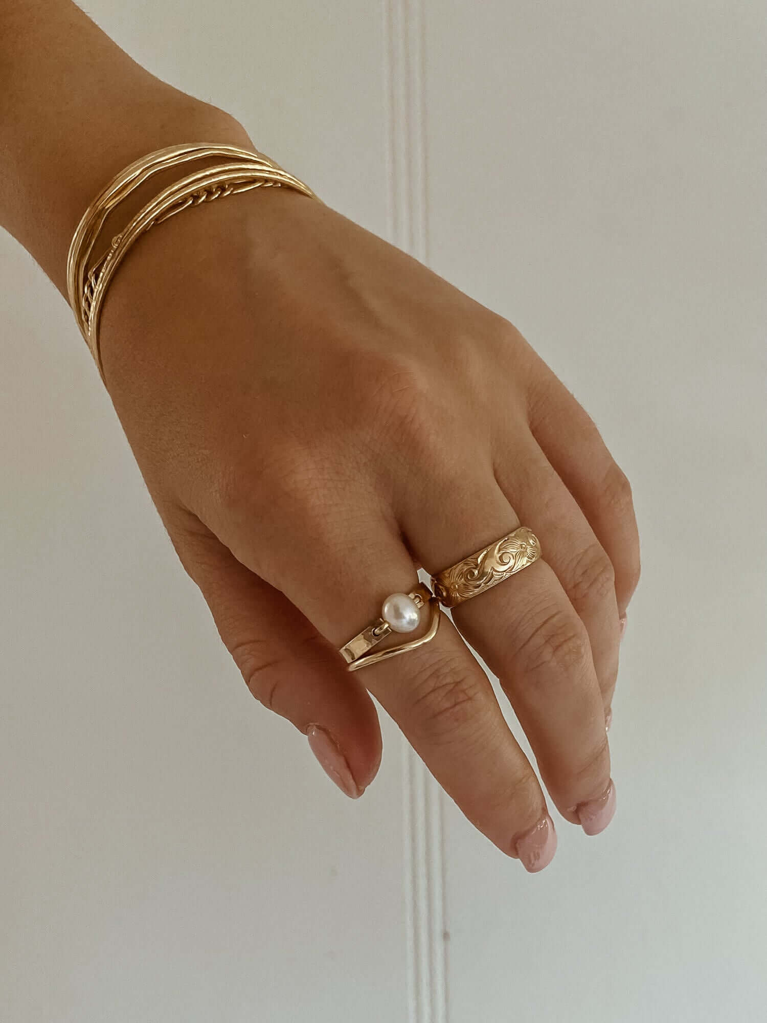 Close-up of a hand adorned with tarnish-proof gold bracelets and two rings, one being the elegant Peak Ring with a pearl, and the other showcasing ornate 14k gold-filled designs.