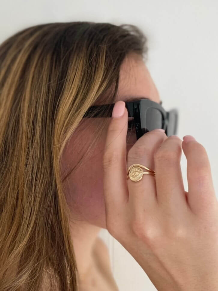 A person with long, light brown hair adjusting black sunglasses, wearing the Peak Ring—a 14k gold filled stacking ring—on their index finger.