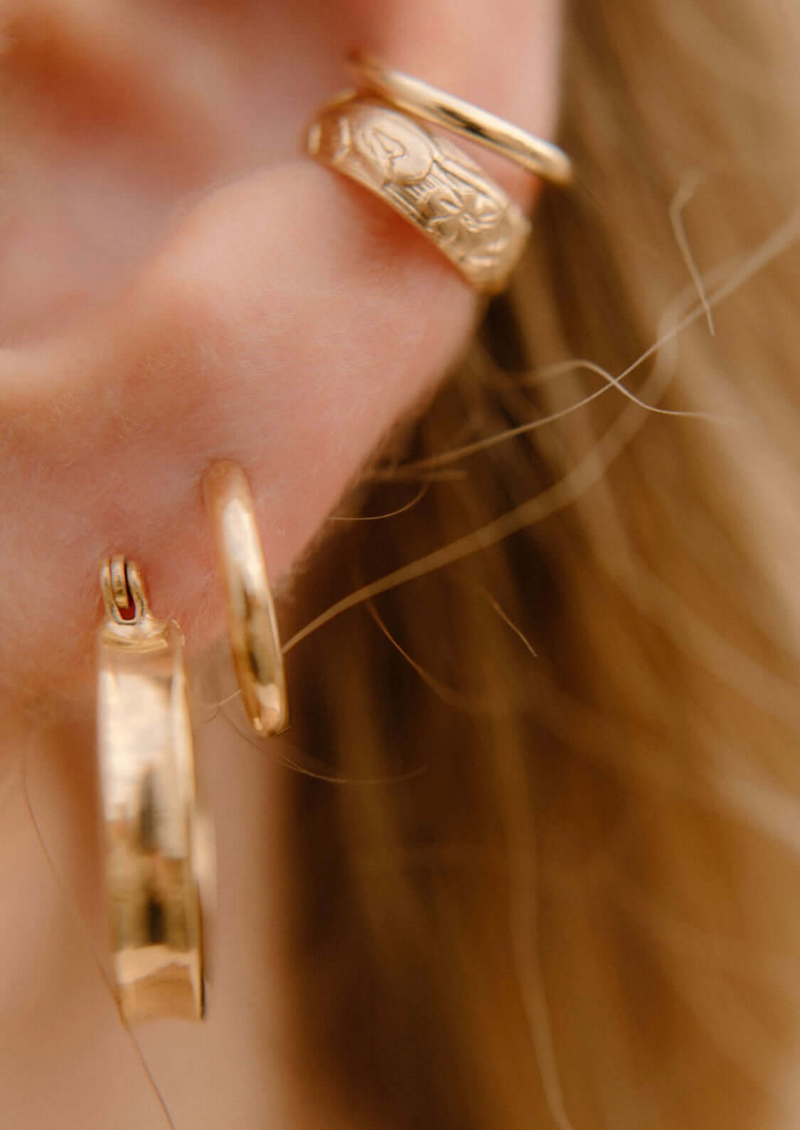 Close-up of a person's ear adorned with three gold hoop earrings, including one with an engraved pattern, alongside the stylish Everyday Ear Cuff.