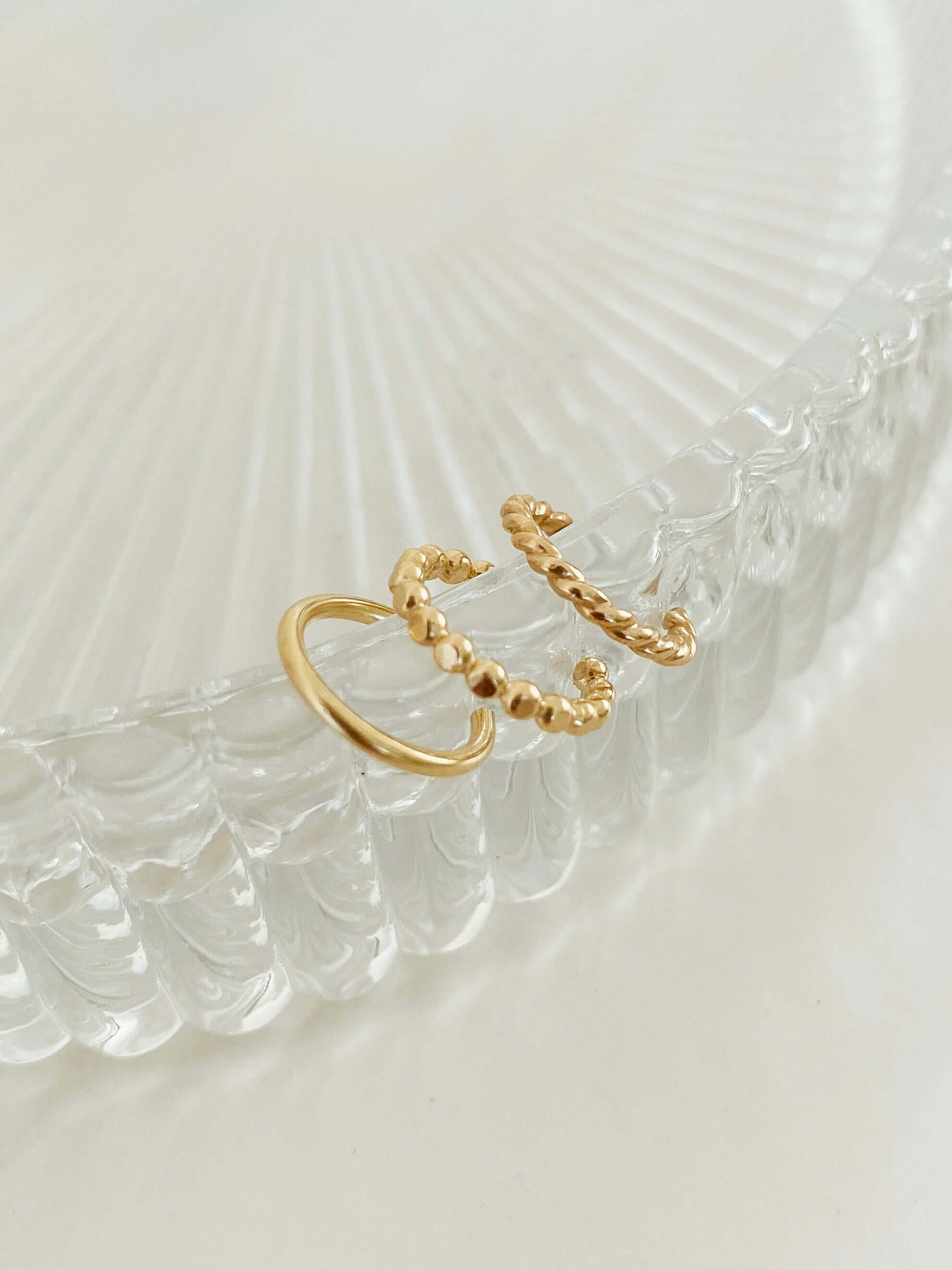 Two gold twisted rings displayed on a clear, ribbed glass surface in soft light embody the elegance of the Triple Ear Cuff Set.