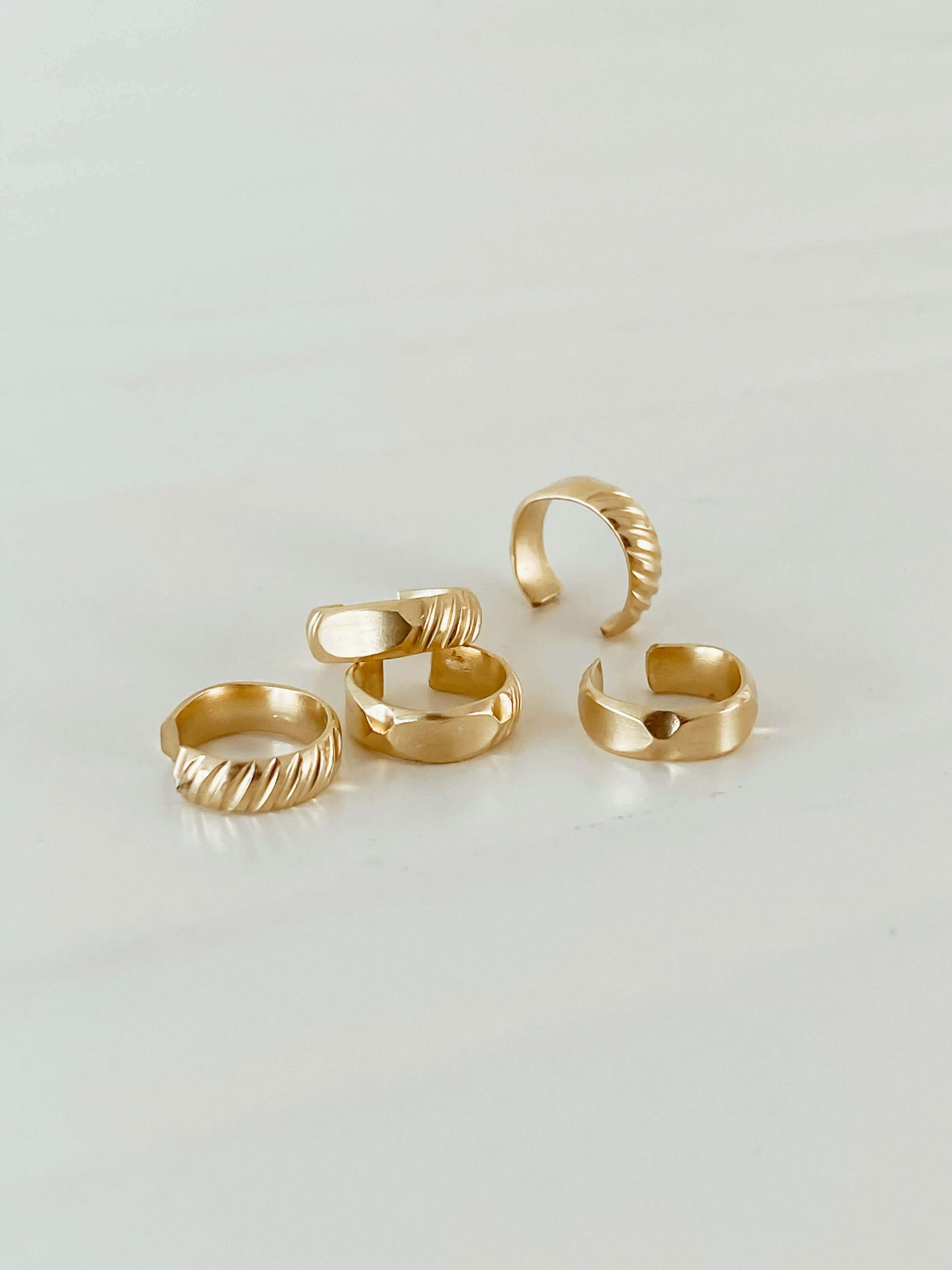 Five gold rings with varied designs displayed on a light surface, each crafted from 14k gold filled material, including the elegant Edge Ear Cuff.