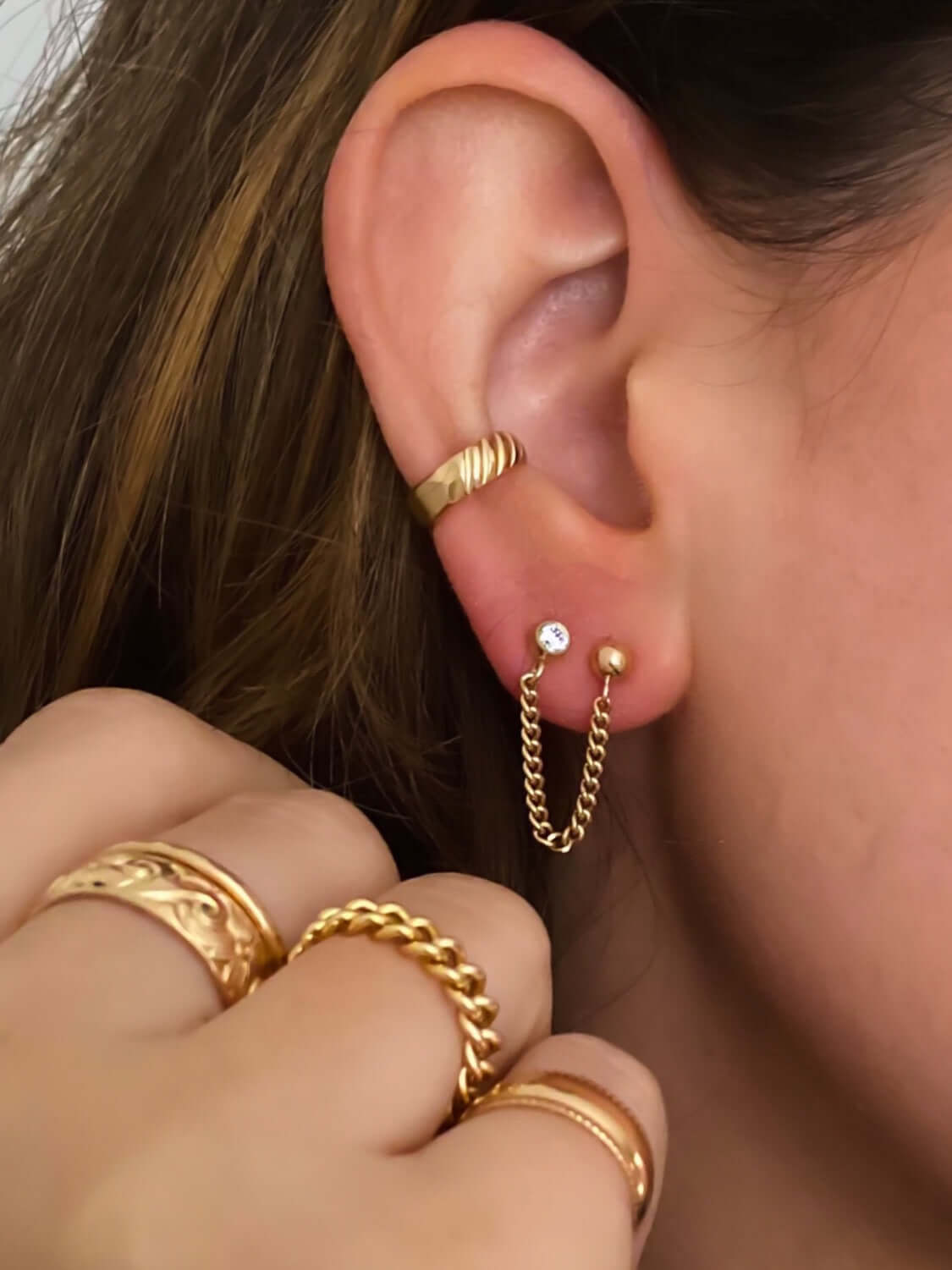 Close-up of an ear adorned with 14k gold filled piercings and a hand showcasing three elegant gold rings. The look is effortlessly chic, with no piercing required thanks to the stylish Edge Ear Cuff.