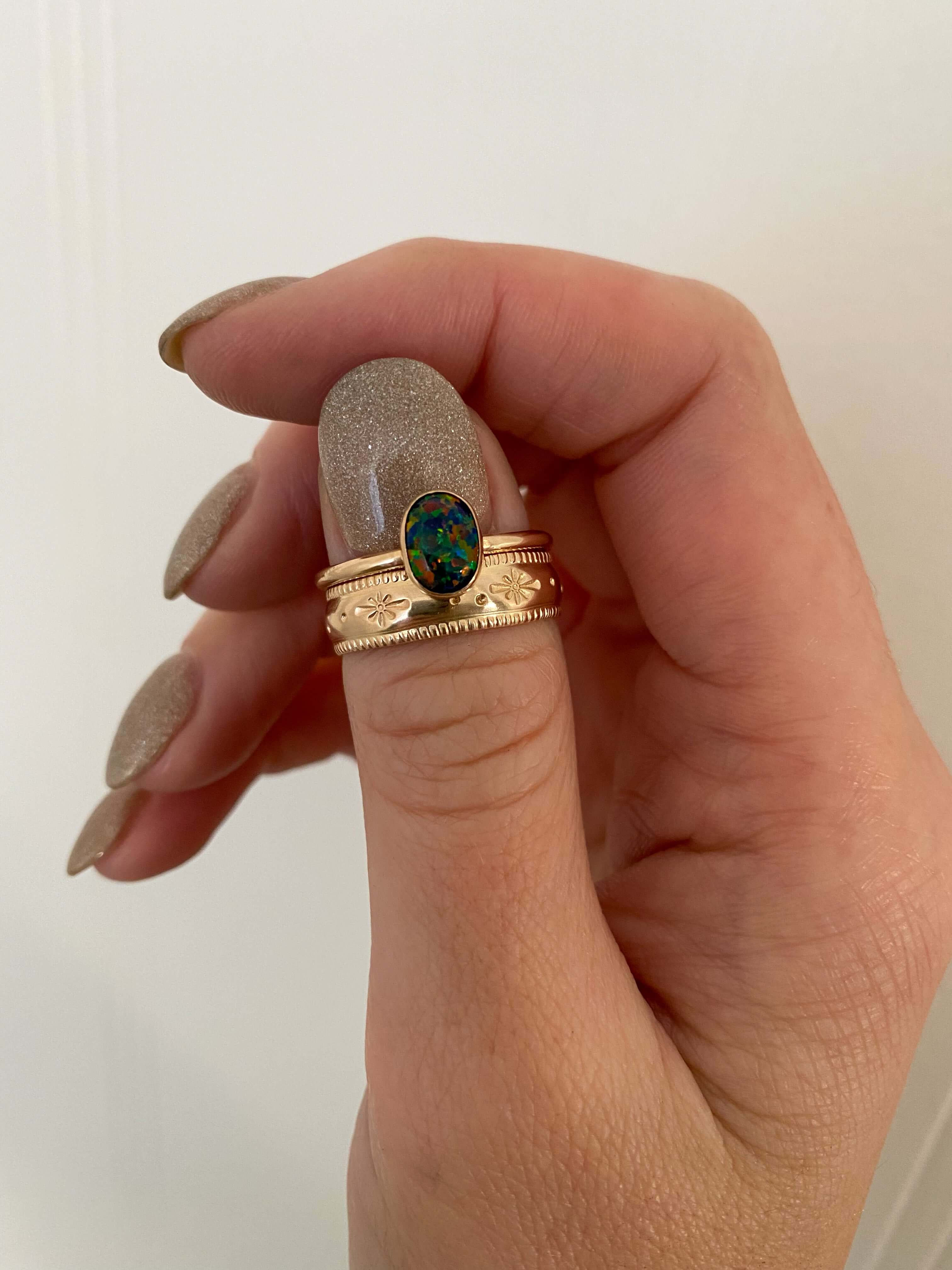 Close-up of a hand with glittery, manicured nails wearing a 14k gold fill Stardust Ring featuring an oval black opal on the thumb.