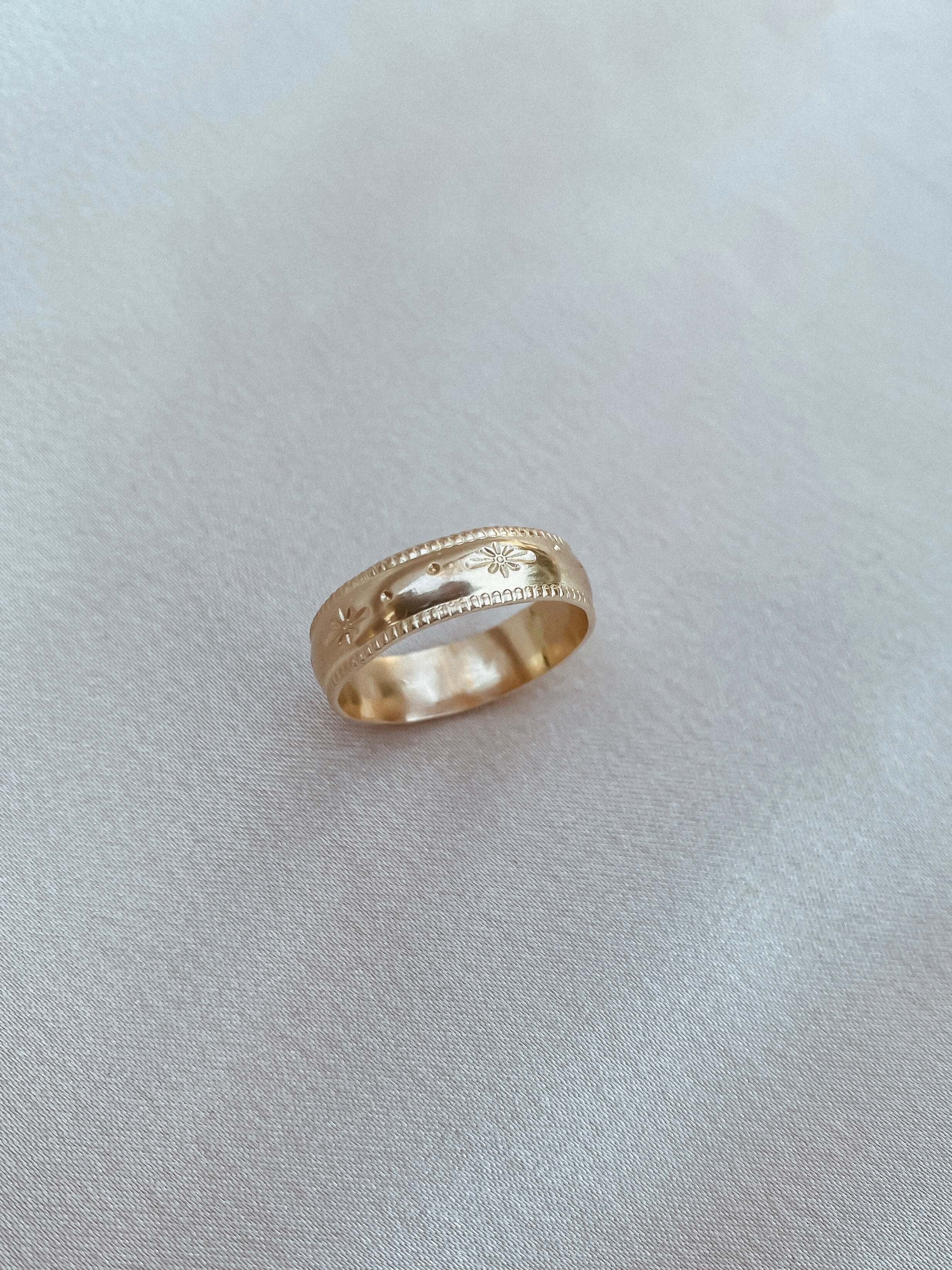 A Stardust Ring, crafted from 14k gold fill and adorned with engraved star and floral patterns, rests gracefully on a light, smooth fabric background, epitomizing celestial jewelry.
