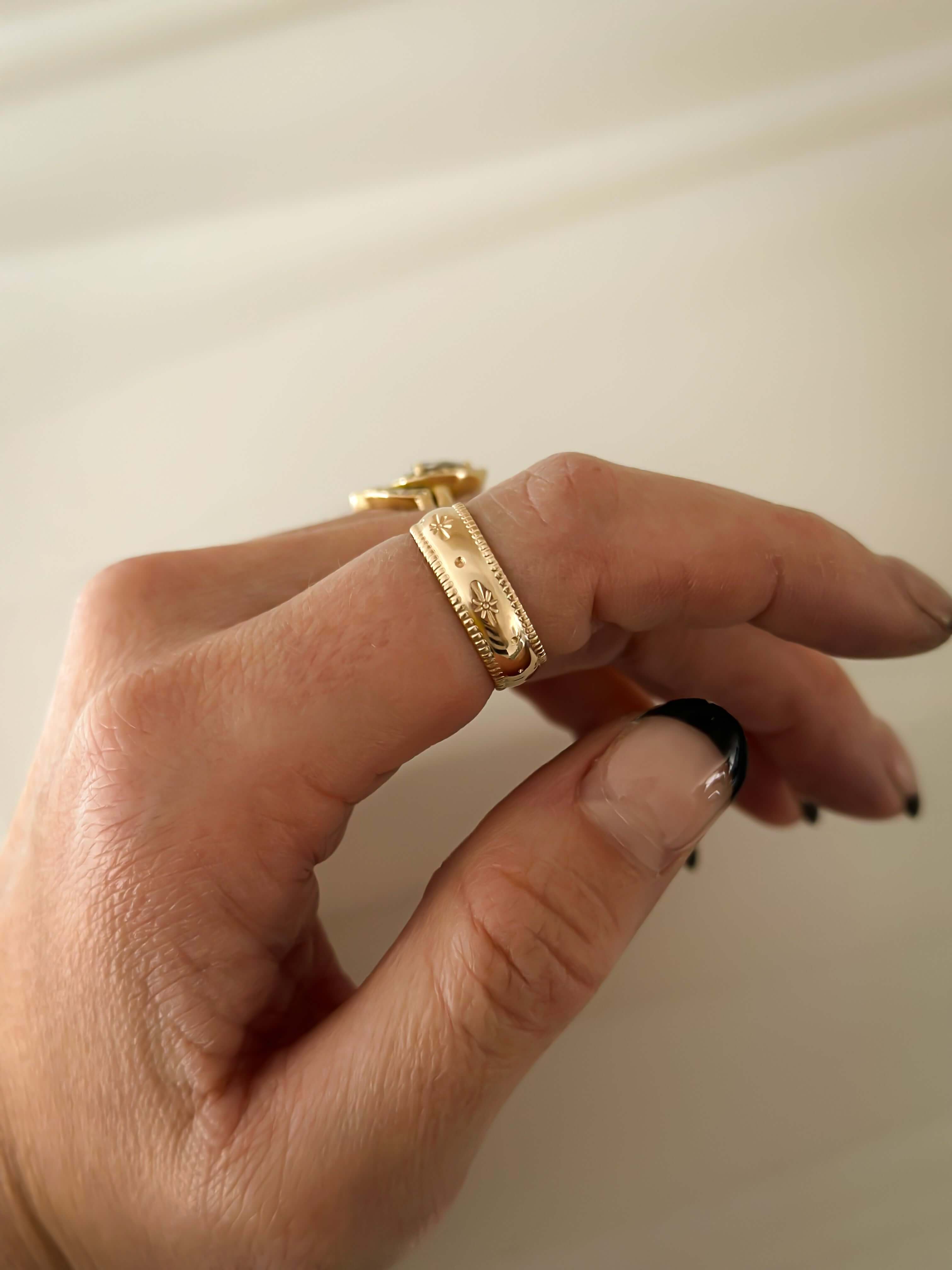 Close-up of a hand wearing the Stardust Ring, a celestial jewelry piece crafted from 14k gold fill with decorative engravings on the middle finger, nails painted in black and beige.