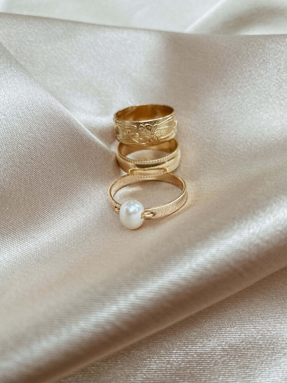 Three gold rings and one Selene Ring adorned with a pearl, elegantly arranged on a silky beige fabric.