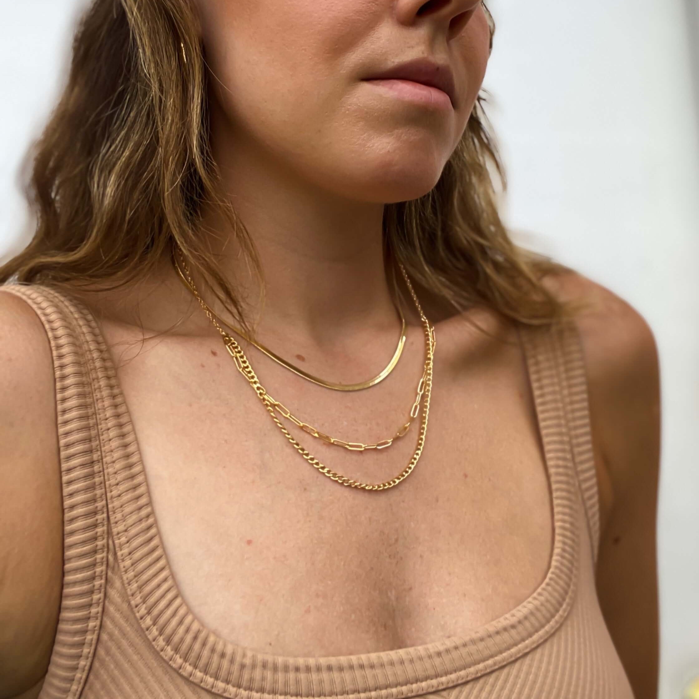 A person with long, wavy hair wears layered gold necklaces, featuring a stylish Selena Chain Necklace, along with a nose ring and a ribbed tank top, with their eyes closed.