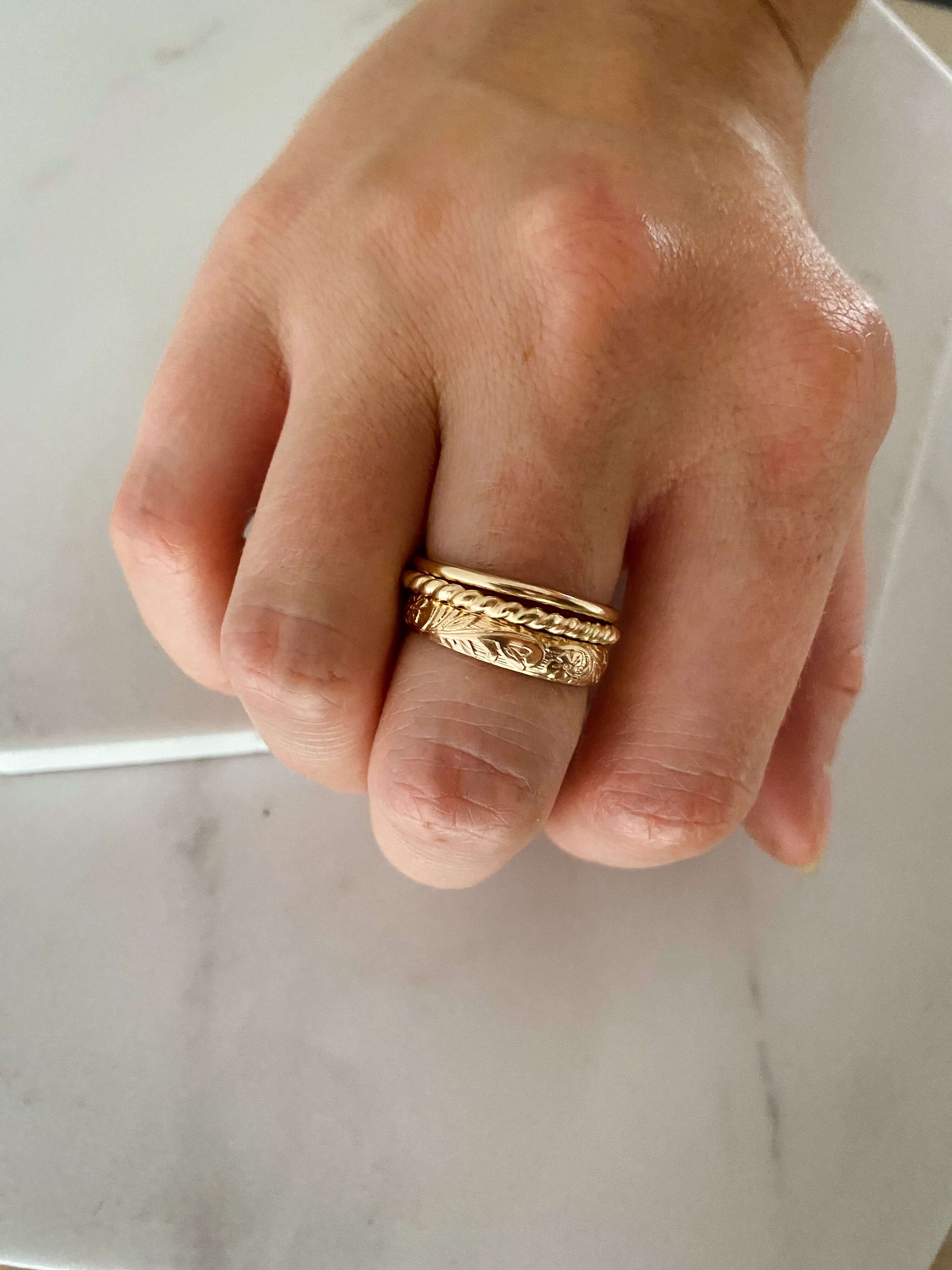 A hand with three gold rings, including the "Simple Gold Band" from our collection, on the ring finger, resting on a white surface.