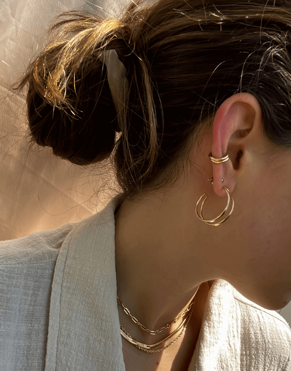 Woman with a bun wearing gold hoop earrings, the Everyday Ear Cuff on her cartilage, a necklace, and a beige blazer, facing left with sunlight in the background.