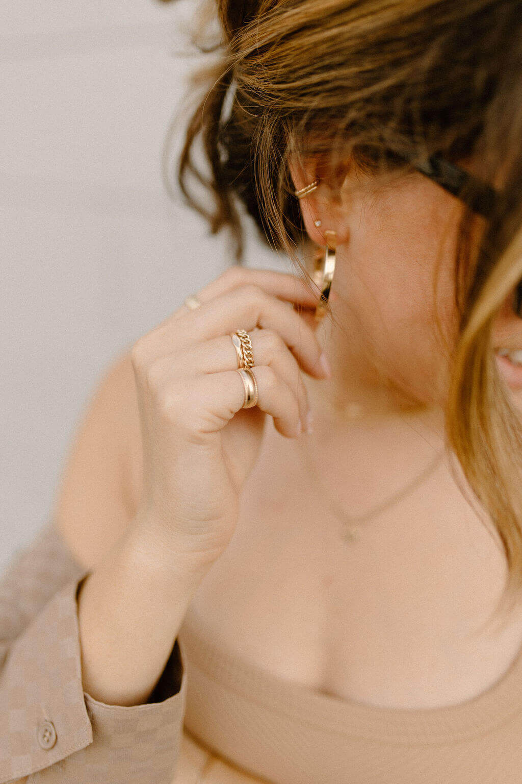Close-up of a woman touching her ear, showcasing her gold rings and earrings. Her wavy brown hair and eyeglasses are visible, and she complements her look with a delicate Selene Ring.