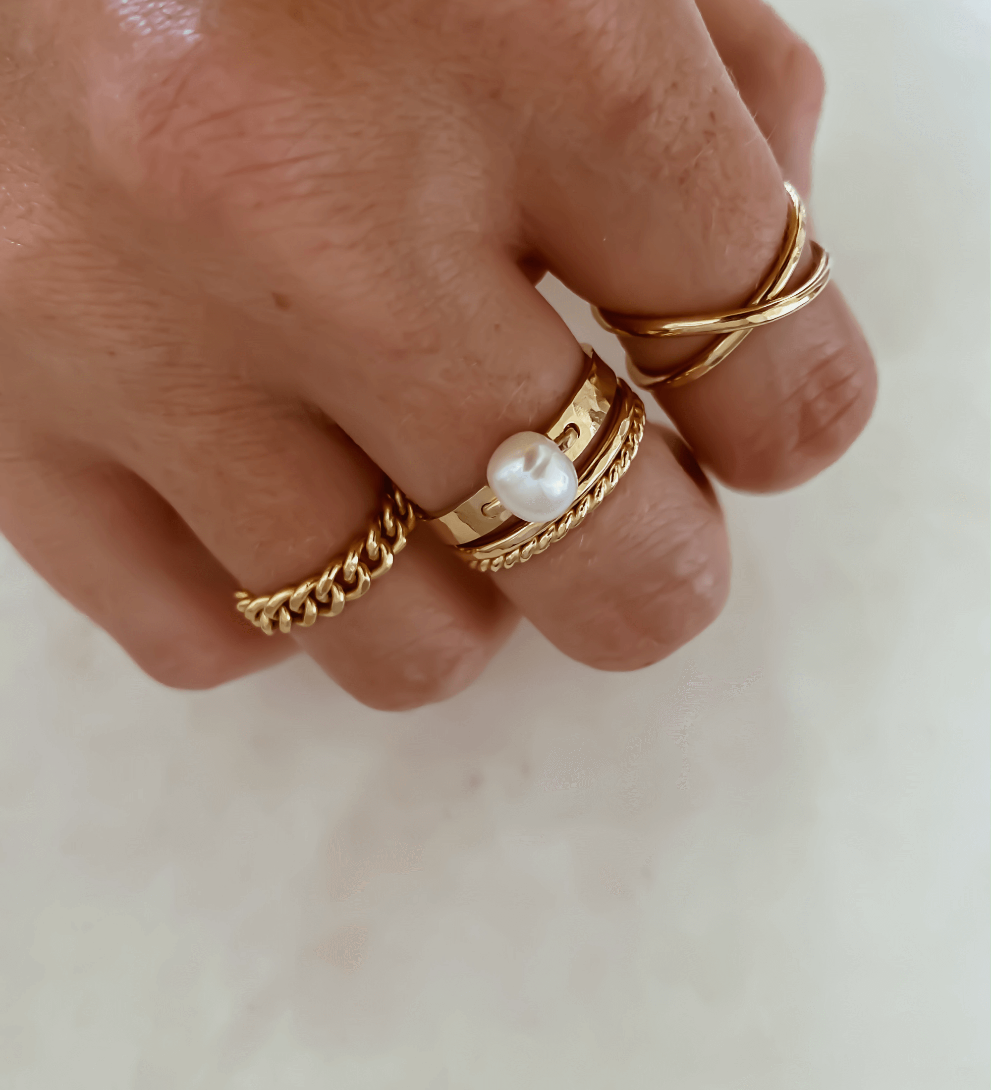 Close-up of a hand adorned with multiple gold rings, including the elegant Thin Twist Band from our collection, a ring with a pearl, and another featuring a chain design, all crafted in 14k gold-filled elegance on a light background.