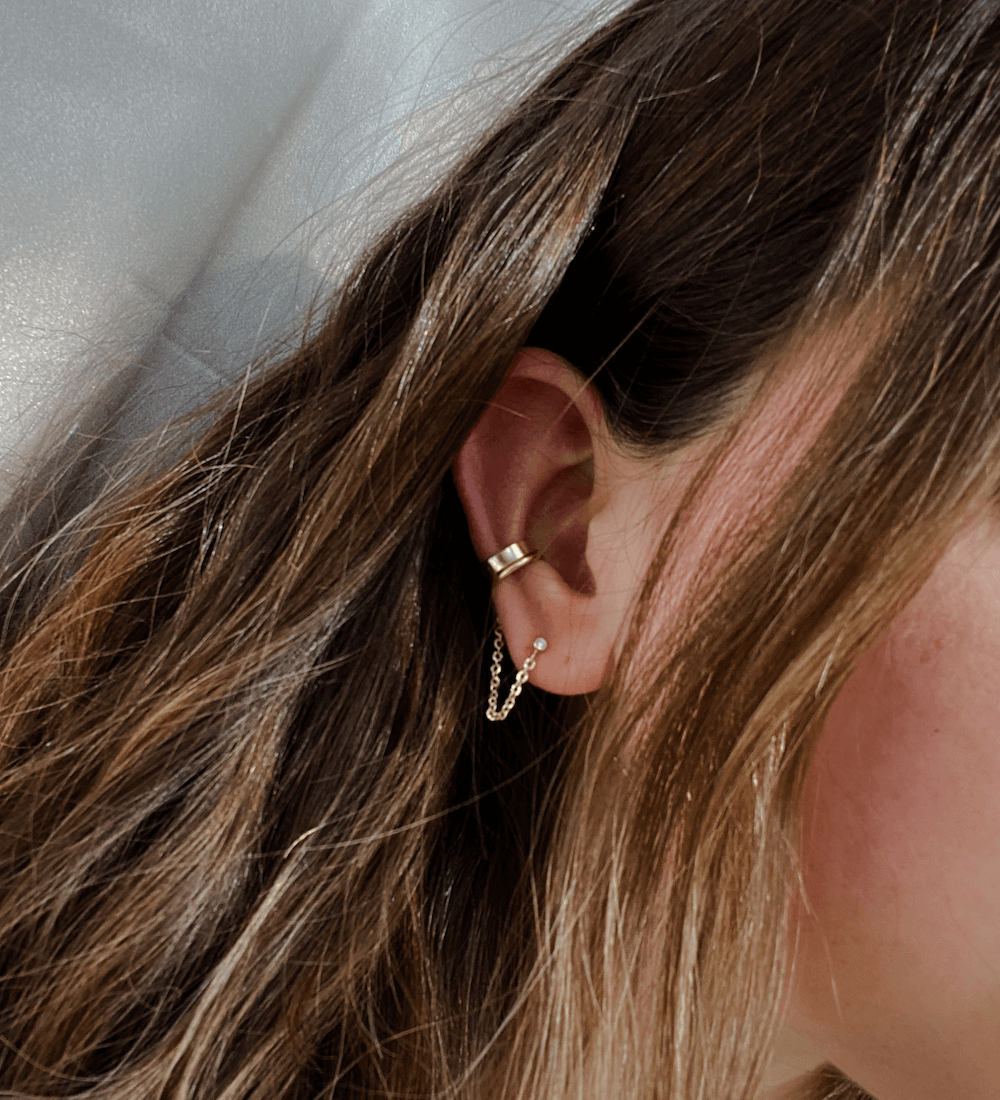 Close-up of a woman's ear with long brown hair, adorned with the polished gold Level Ear Cuff and a chain earring.