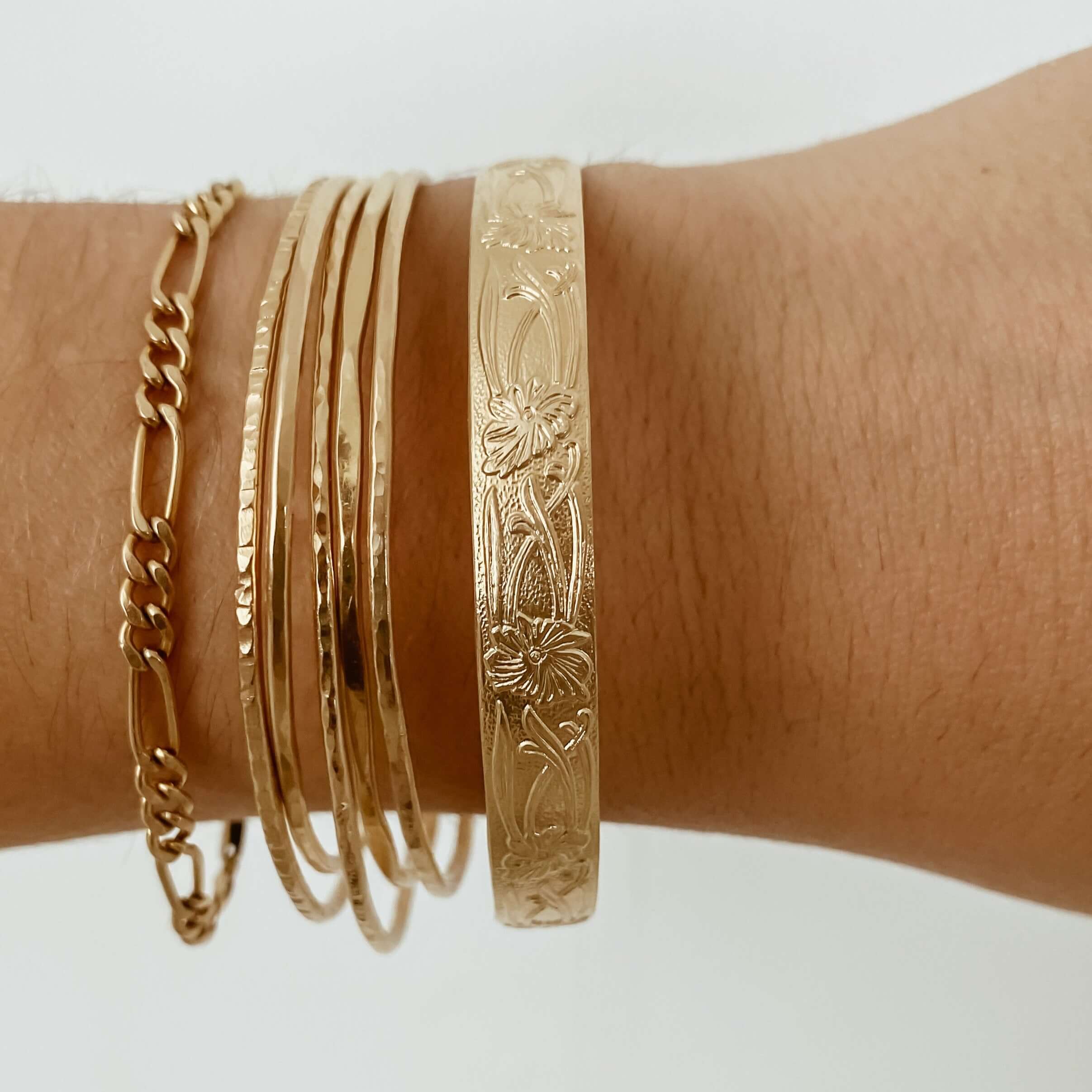 A wrist adorned with multiple gold bangles, including the intricately designed Marigold Cuff Bracelet featuring beautiful floral patterns.