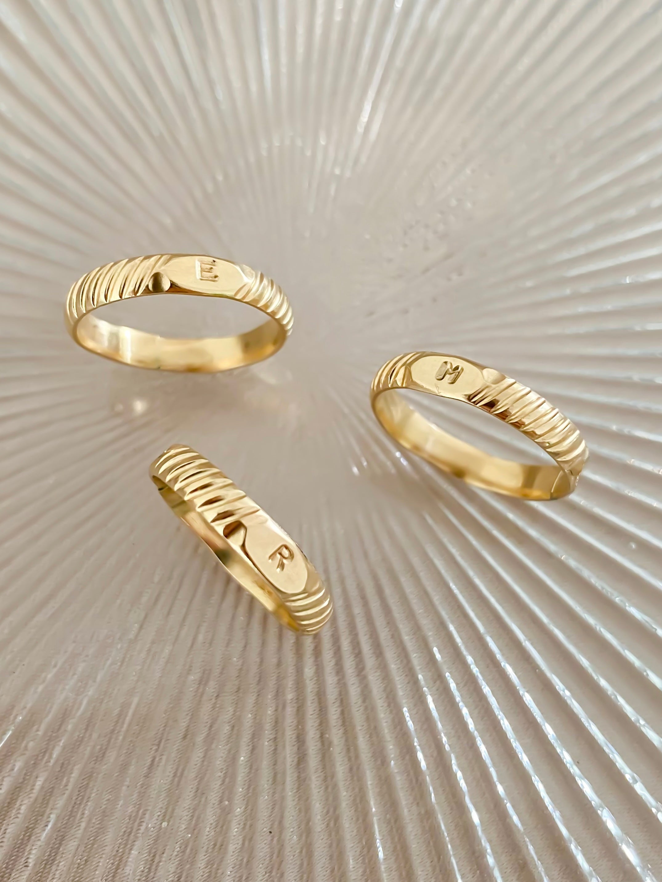 Three gold rings with engraved letters "E," "M," and "R" are displayed on a textured, radial pattern surface.