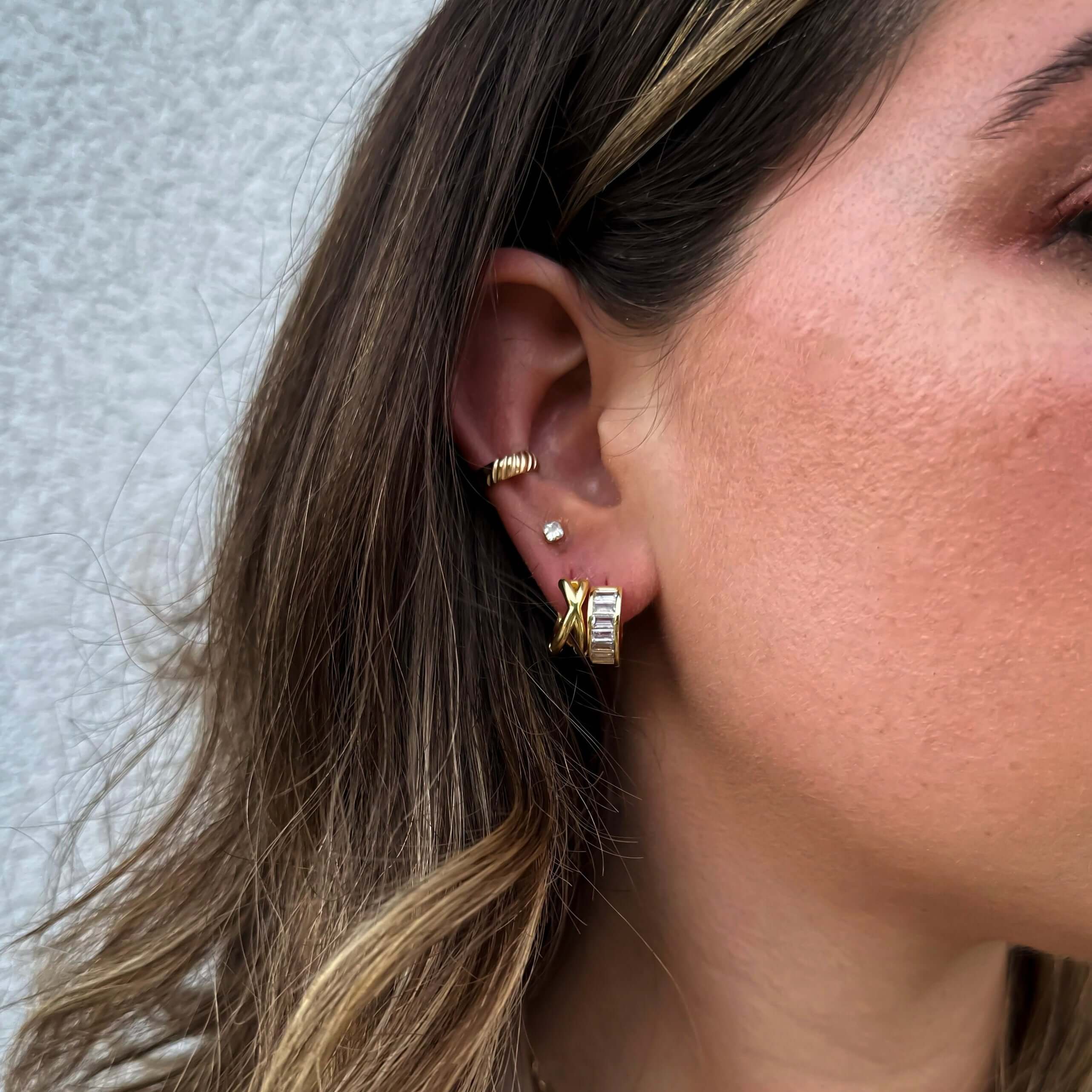 Close-up of a woman's ear adorned with multiple 18k gold plated, tarnish-proof Baguette Hoop Earrings and diamond studs against a light background.