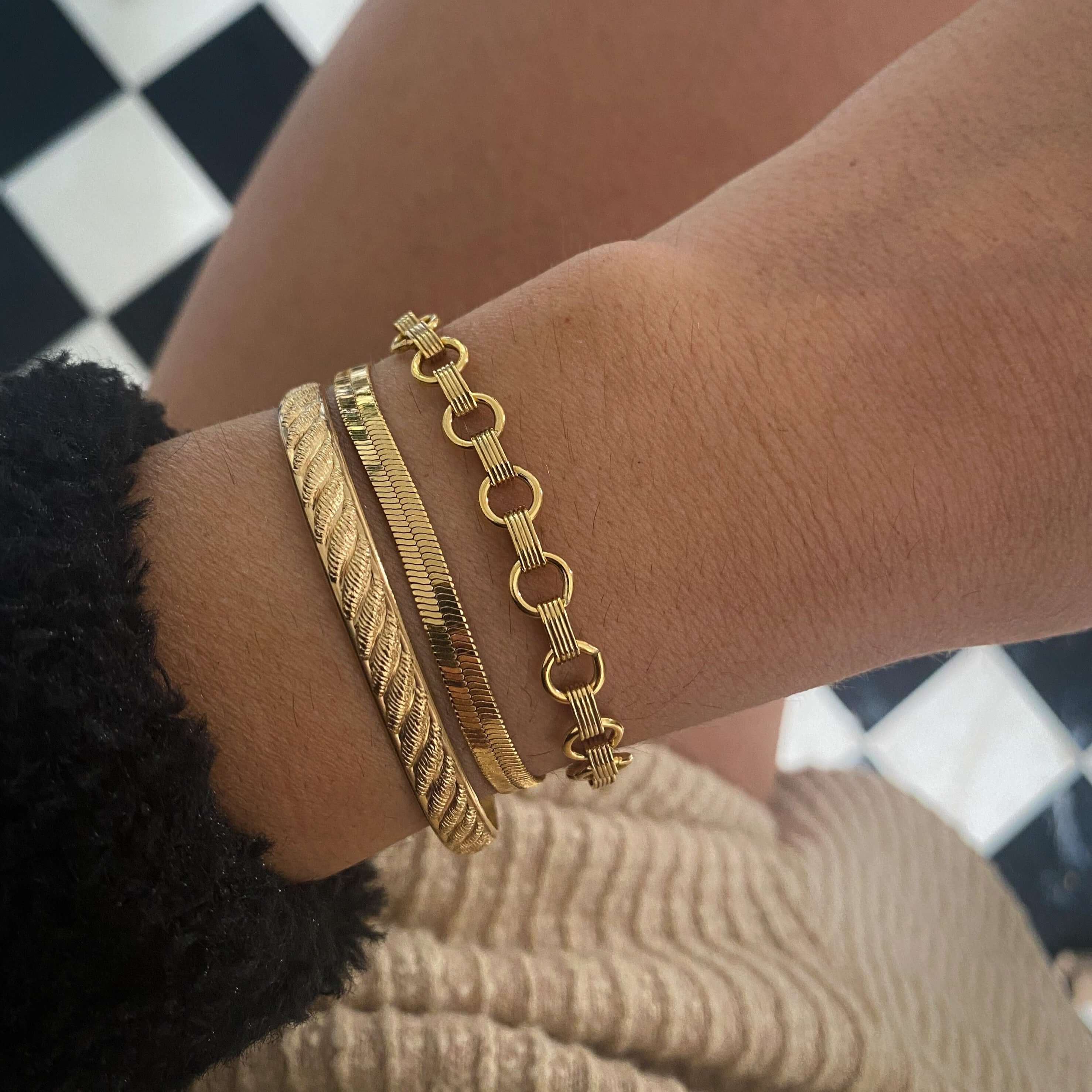 A wrist adorned with three Bold Herringbone Bracelets, each crafted in exquisite 18K gold and showcasing unique designs, stands out against a checkered black and white tile background.