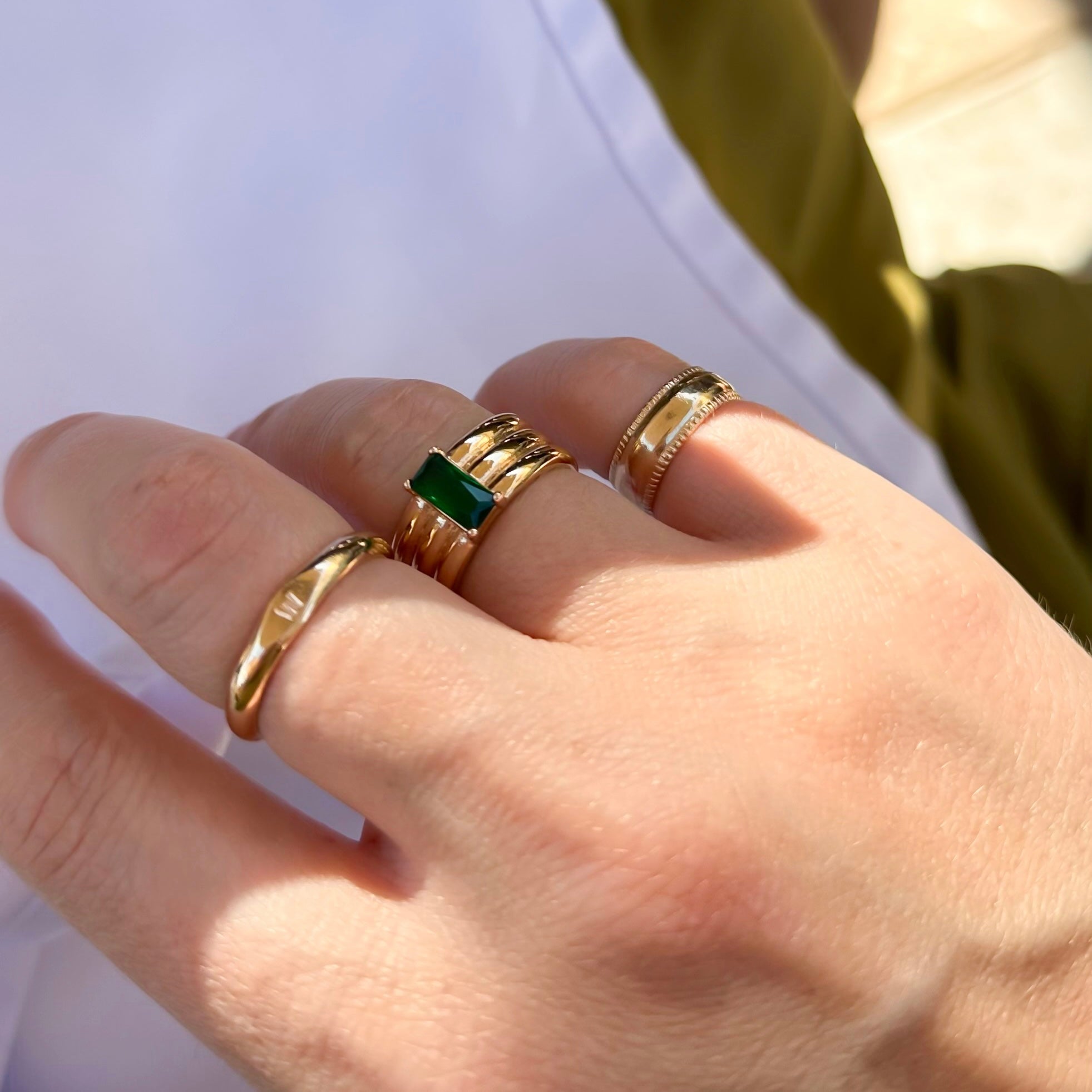 A hand adorned with multiple gold rings, including the stunning Envy Emerald Ring featuring a green gemstone, is showcased against a blurred background. This wearable masterpiece highlights intricate designs, elevating any outfit to new heights.