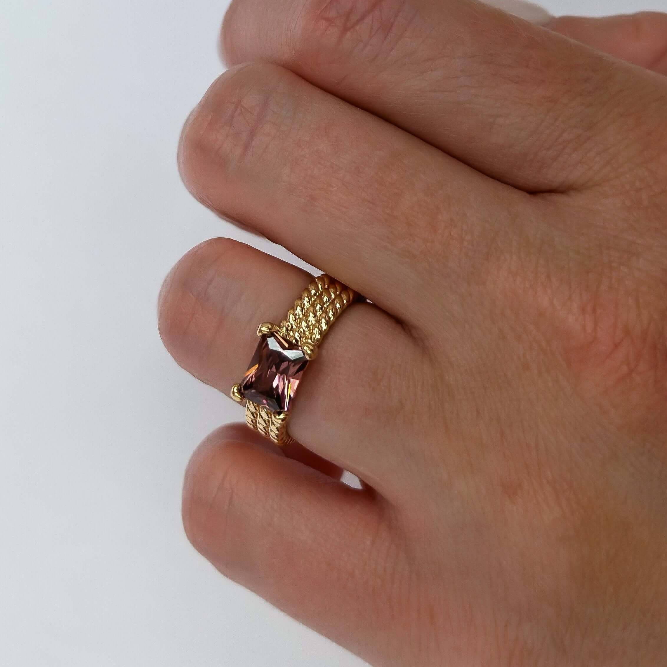 Close-up of a hand wearing the Ivy Ring - Magenta, featuring a rectangular magenta gemstone on a gold band, adorning the ring finger against a white background and radiating an air of regal jewelry.