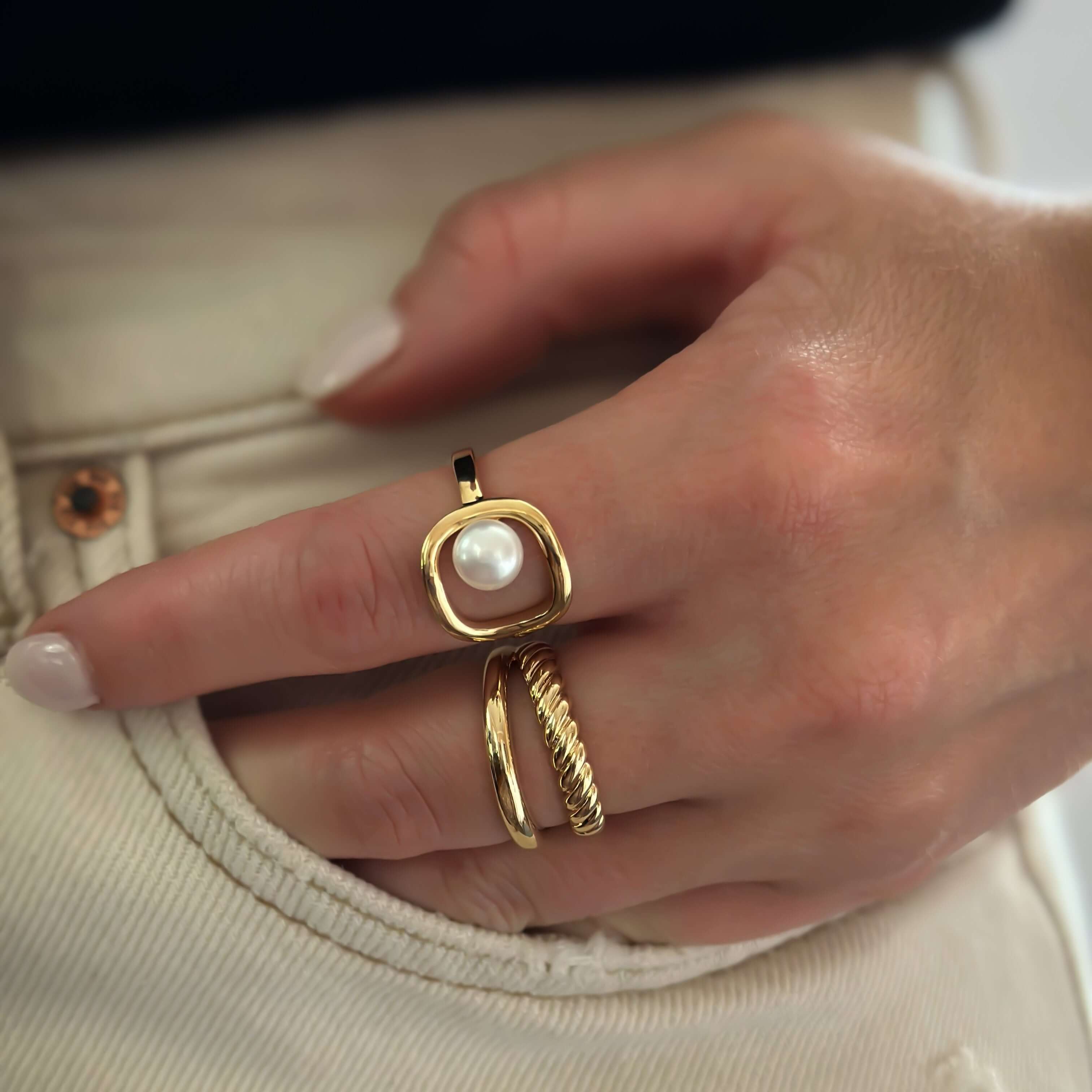 A hand adorned with stylish gold rings, including the Plaza Pearl Ring featuring a cultured pearl and unique design, rests against beige pants with a copper button.