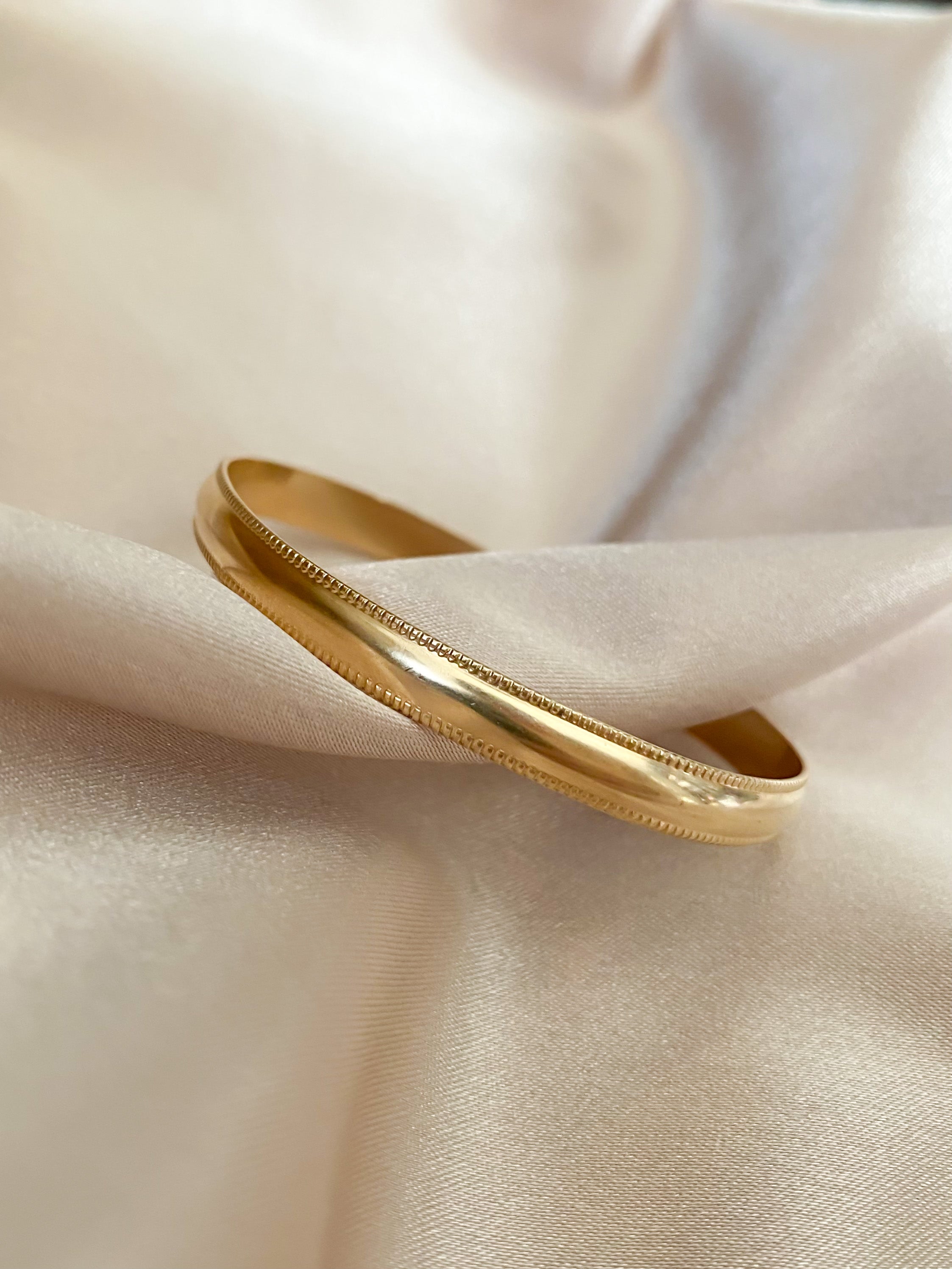 A gold bangle bracelet with intricate detailing, placed on silky beige fabric.